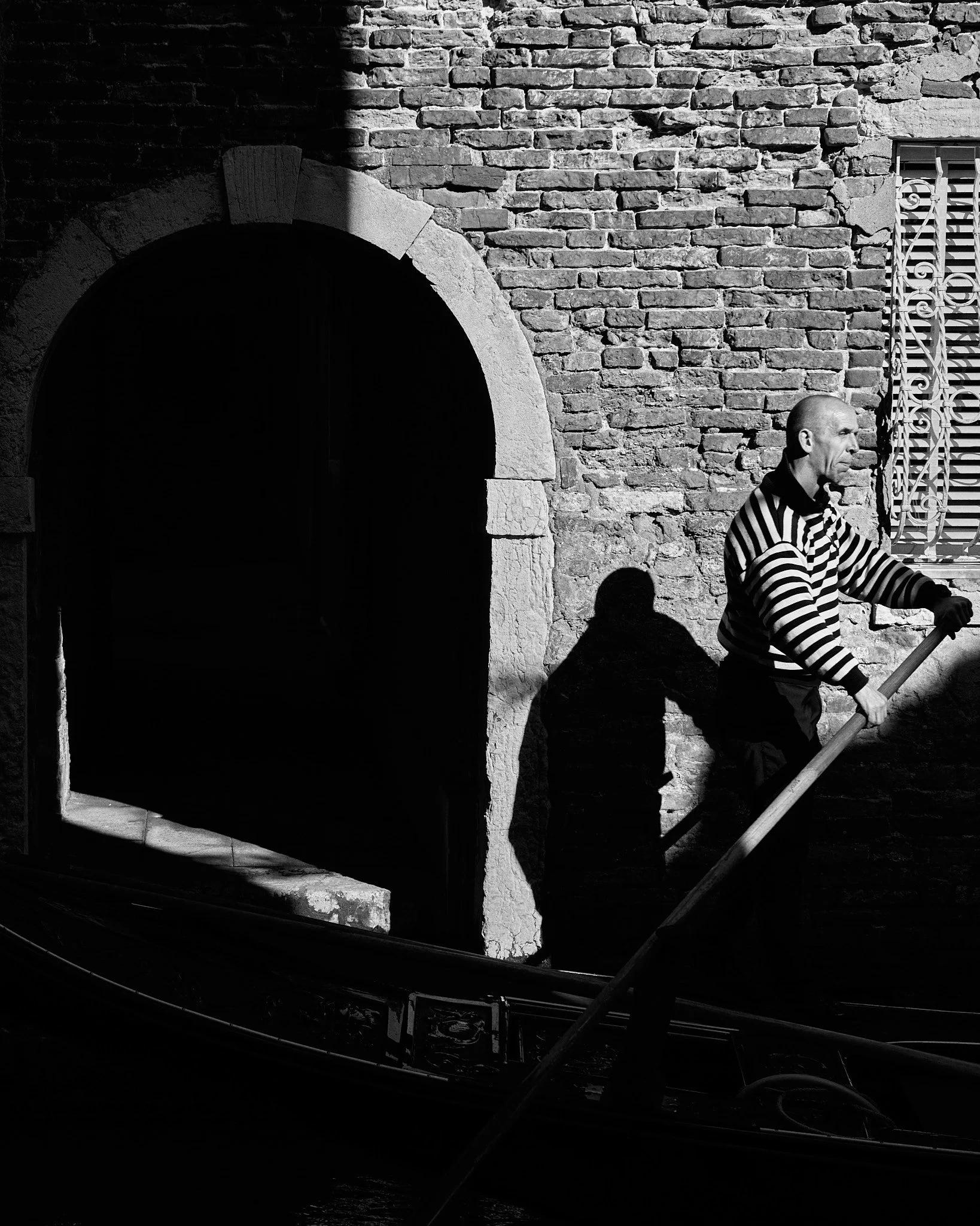 A man with a shaved head and striped shirt pushes a boat along a canal, with a brick building and a window with decorative metal bars in the background. The sunlight casts a shadow of the man on the brick wall.