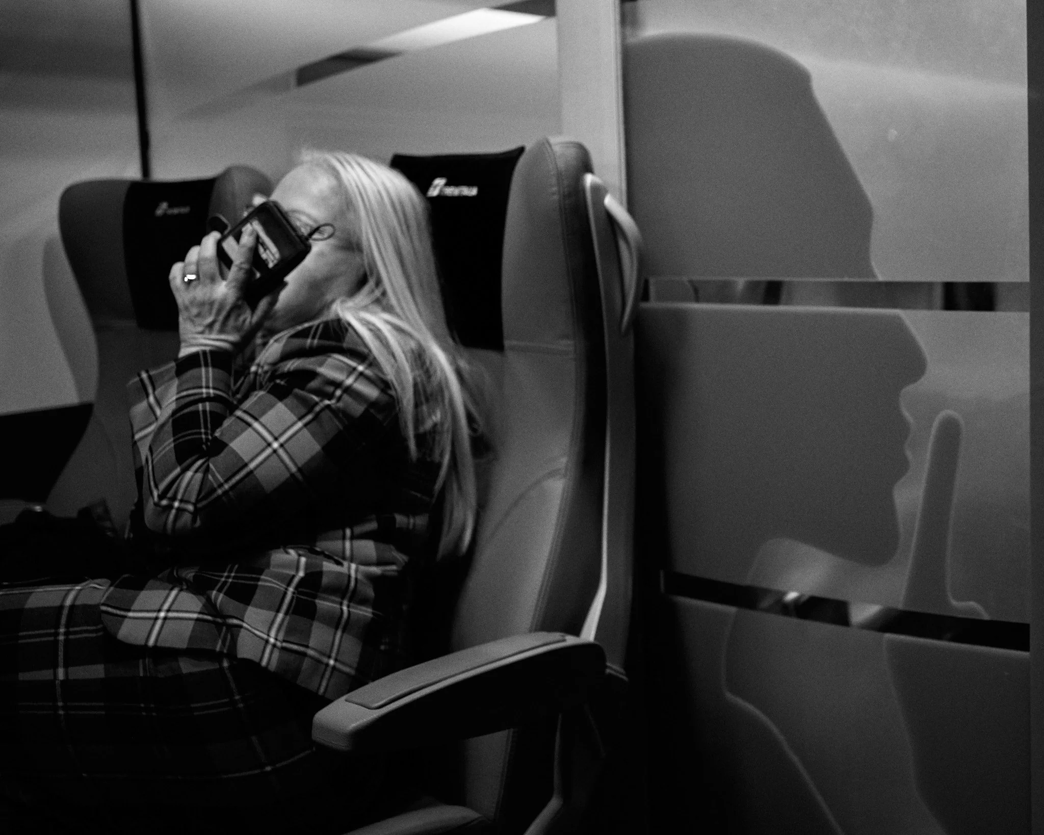 A woman with long hair and a plaid shirt sitting in an airplane seat, talking on a phone, with her reflection visible on a window or partition to her right.