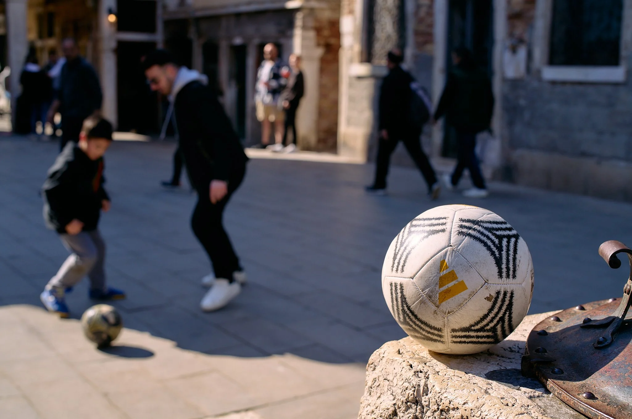 A soccer ball resting on a stone ledge in a historic European city street, with people and buildings in the background, during daytime.