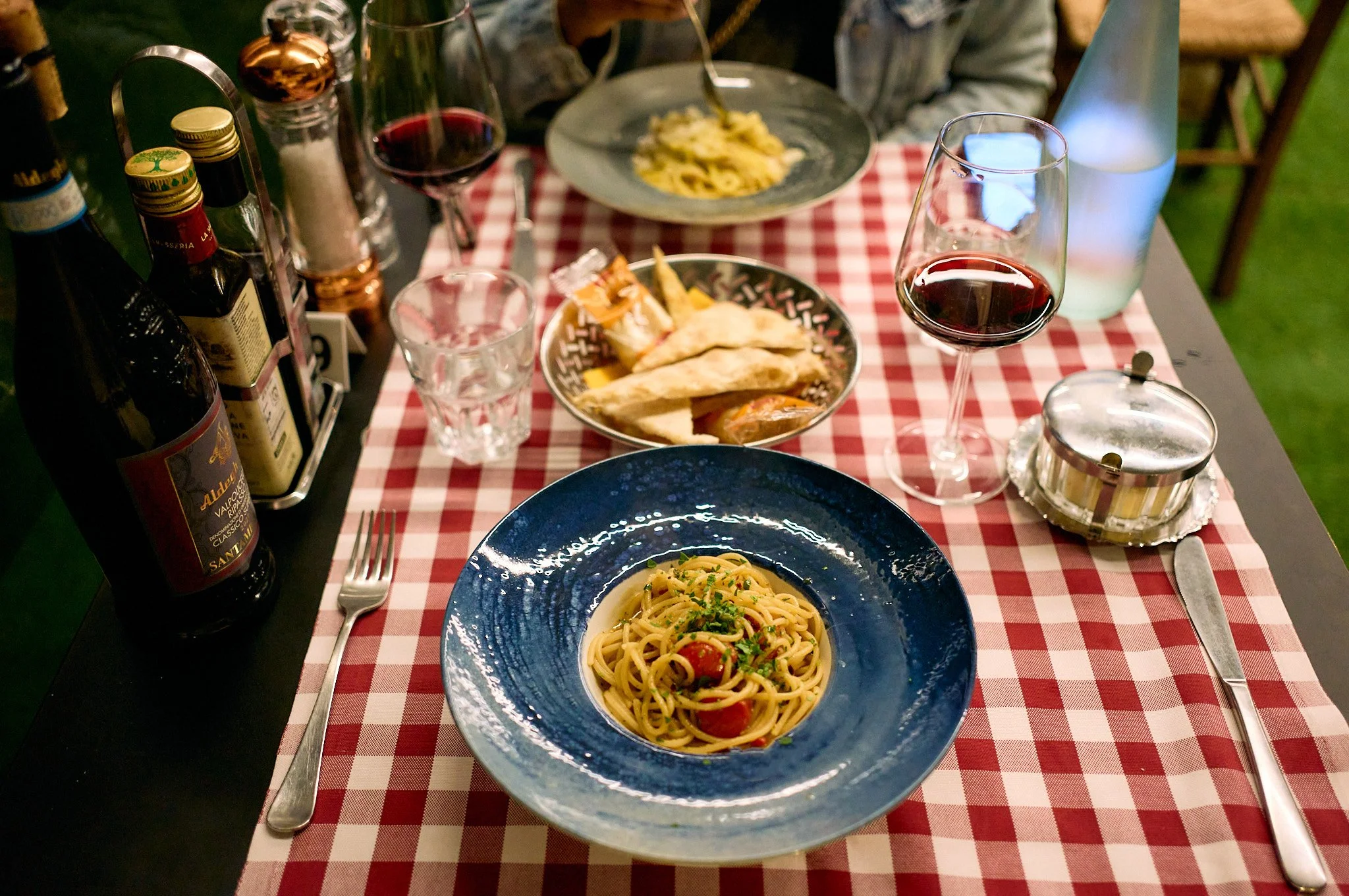 A dinner table with a bowl of spaghetti and tomato sauce, a dish of assorted bread and cheese, red wine, water, and condiments on a red and white checkered tablecloth.