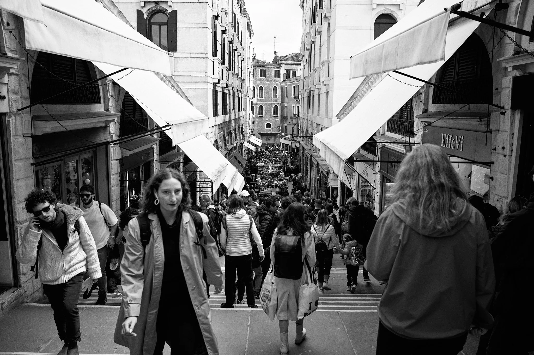 Crowded city street with people walking down steps between buildings lined with shops and cafes; awnings over storefronts.