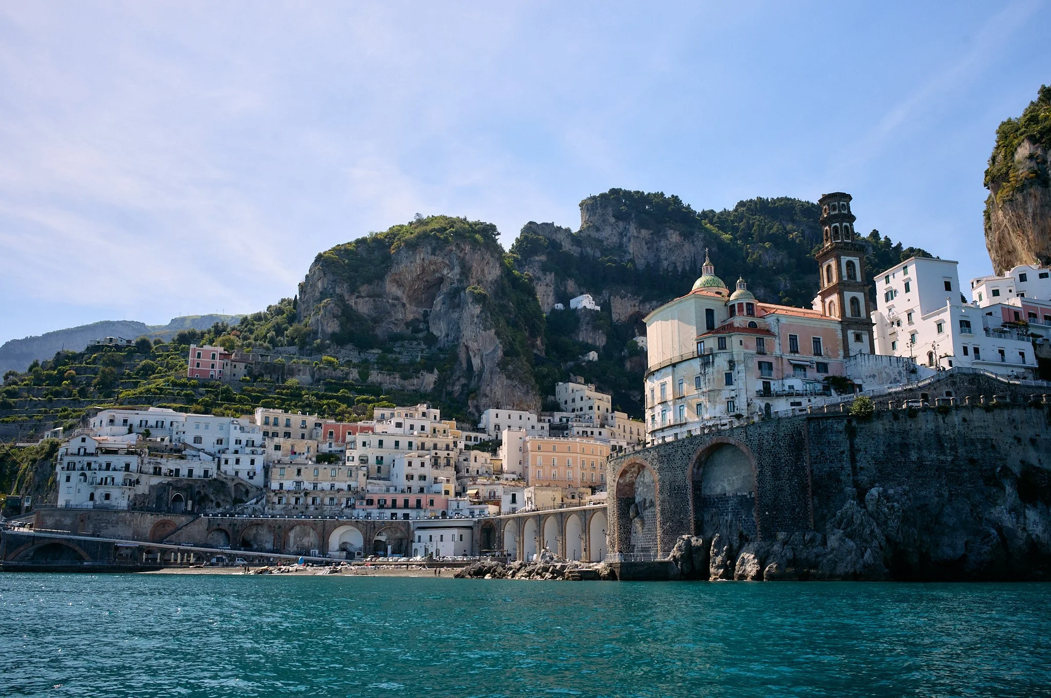 Coastal view of a hillside village with white and pastel-colored buildings, a church with a dome and bell tower, and cliffs in the background, along a turquoise water shoreline under a clear blue sky.
