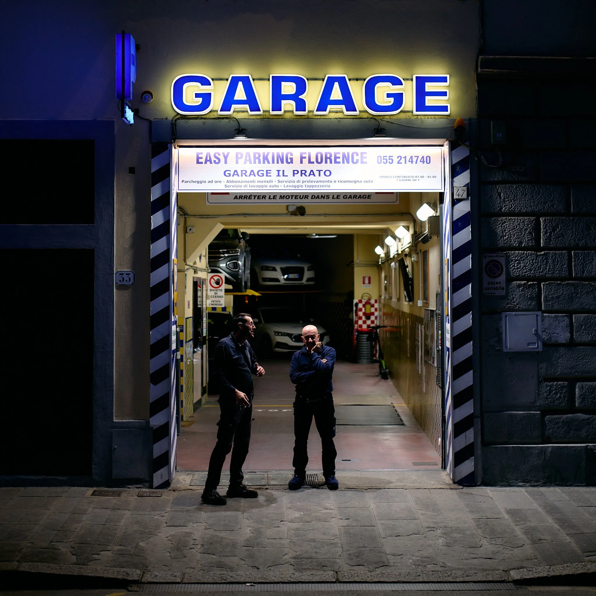 Night scene of an open car garage with two men standing outside, illuminated by bright lights. Inside the garage, there are cars parked and some equipment visible.