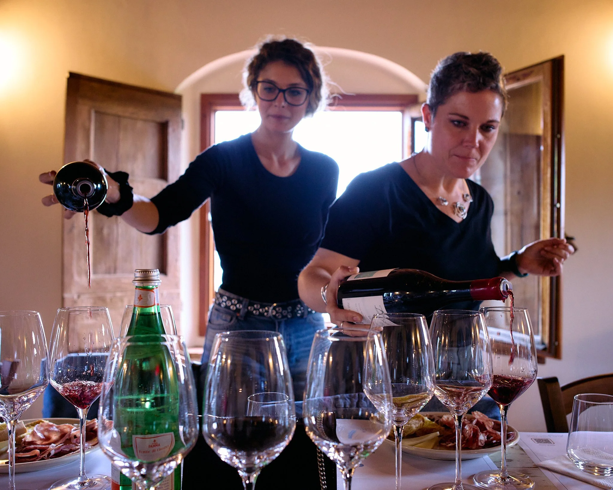Two women serving wine at a table with multiple wine glasses and plates of food in a warmly lit room.