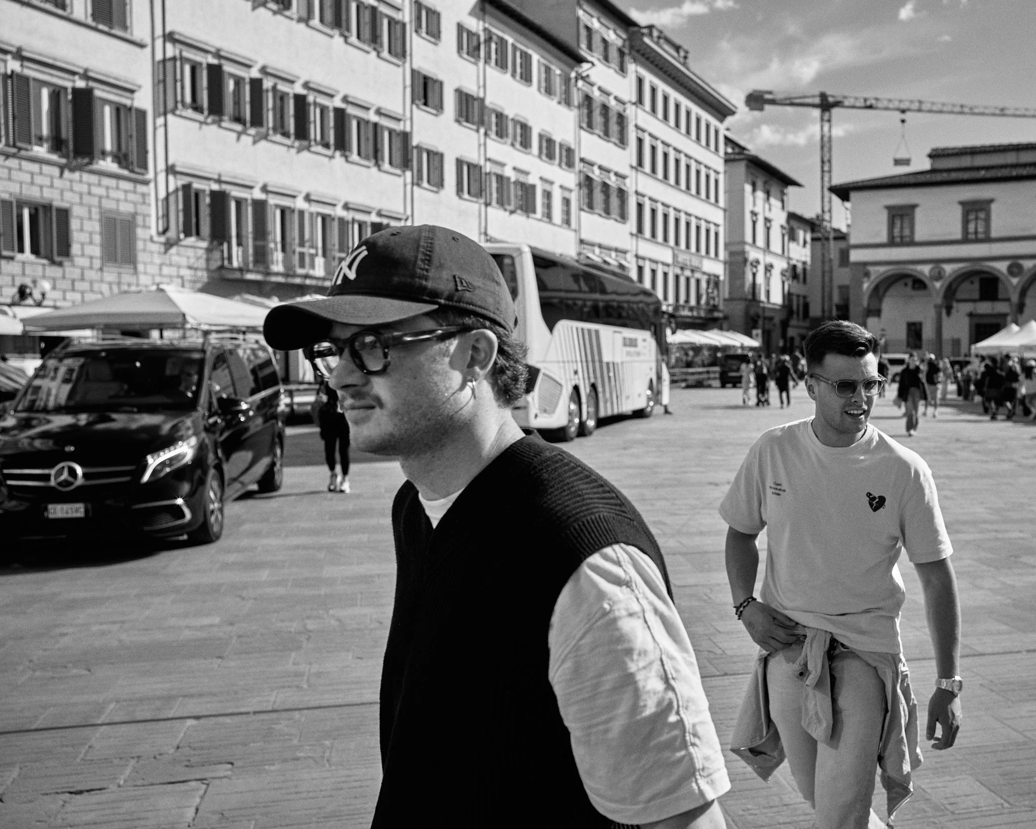 Two young men walking in a city square with historic buildings, cars, and a construction crane in the background. One man is wearing a cap, glasses, and a sweater vest, while the other is wearing sunglasses and a T-shirt.