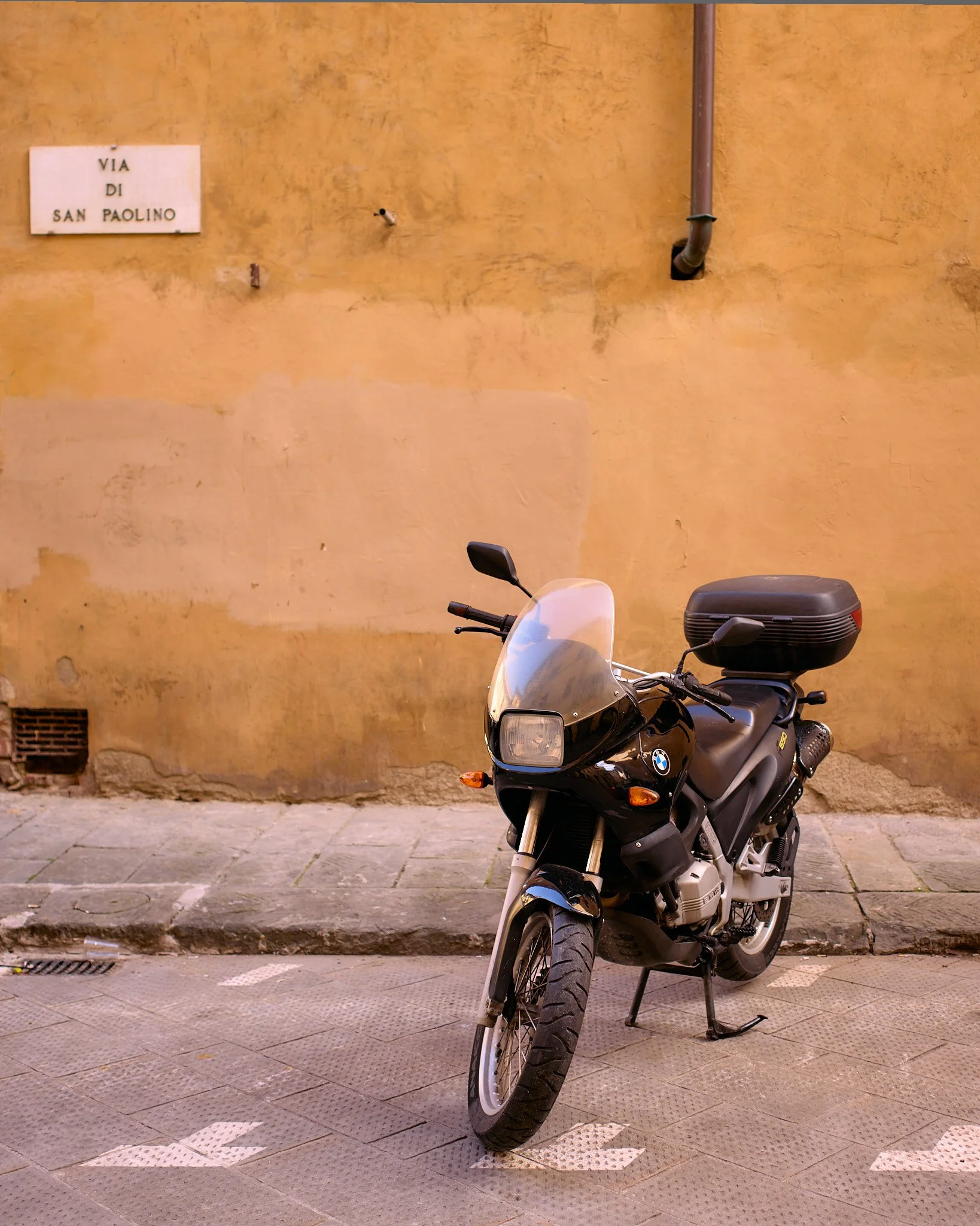 A black BMW motorcycle parked on a cobblestone street next to a yellow stucco wall with a street sign that reads 'VIA DI SAN PAOLINO'.