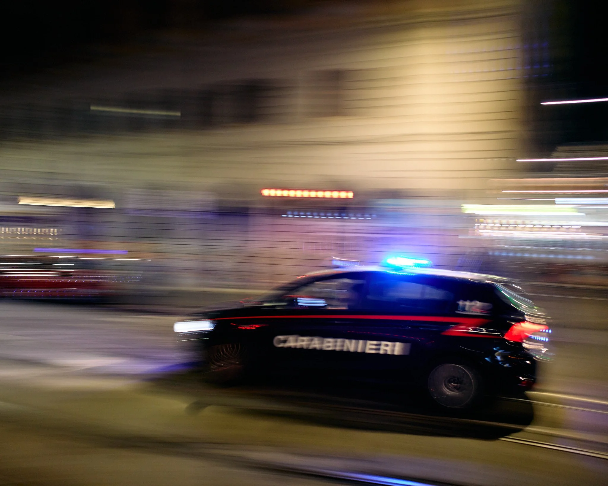 A blurred motion photo of a black police car with flashing blue lights and the word 'Carabinieri' on the side, against a city street background at night.