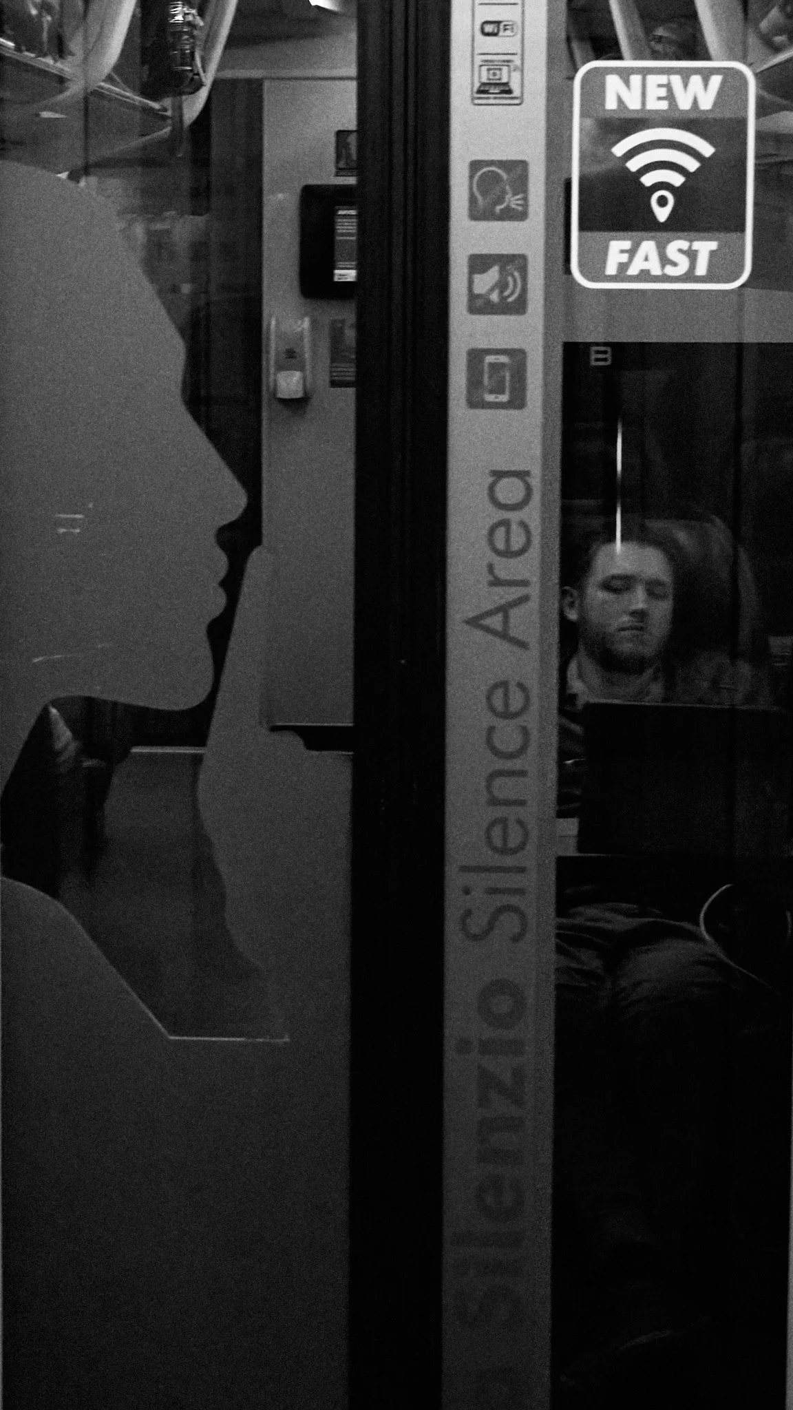 A person appears to be taking a photo reflected in the window of a business. Inside, a man is sitting at a table with a laptop. The window features stickers, including a sign reading 'Decision, Silence, Area' vertically and a sticker advertising 'New