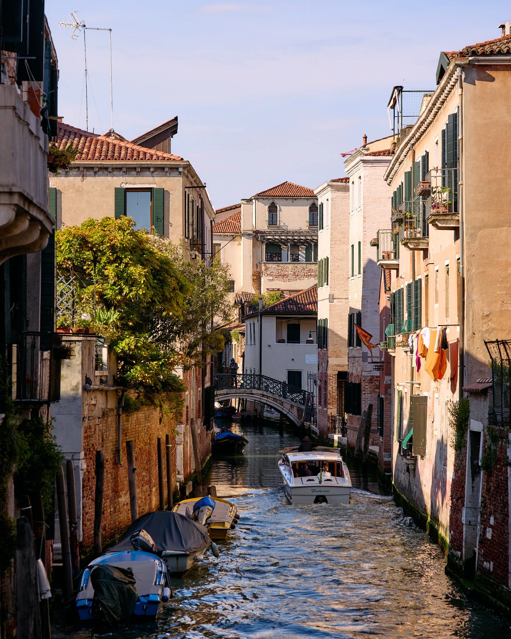 A canal in Venice, Italy, with boats floating and a small bridge connecting the buildings on either side. Colorful old buildings with green shutters line the canal, some with clothes hanging on the balconies. A tree with green foliage is on the left 