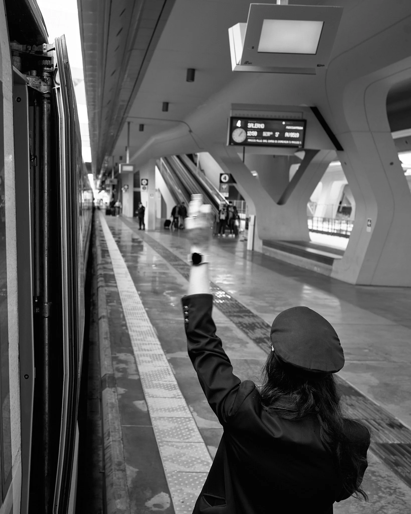 A woman in a dark jacket and hat tossing a can into a recycling bin at a modern train station. The station features escalators, digital information displays, and a few passengers in the background.