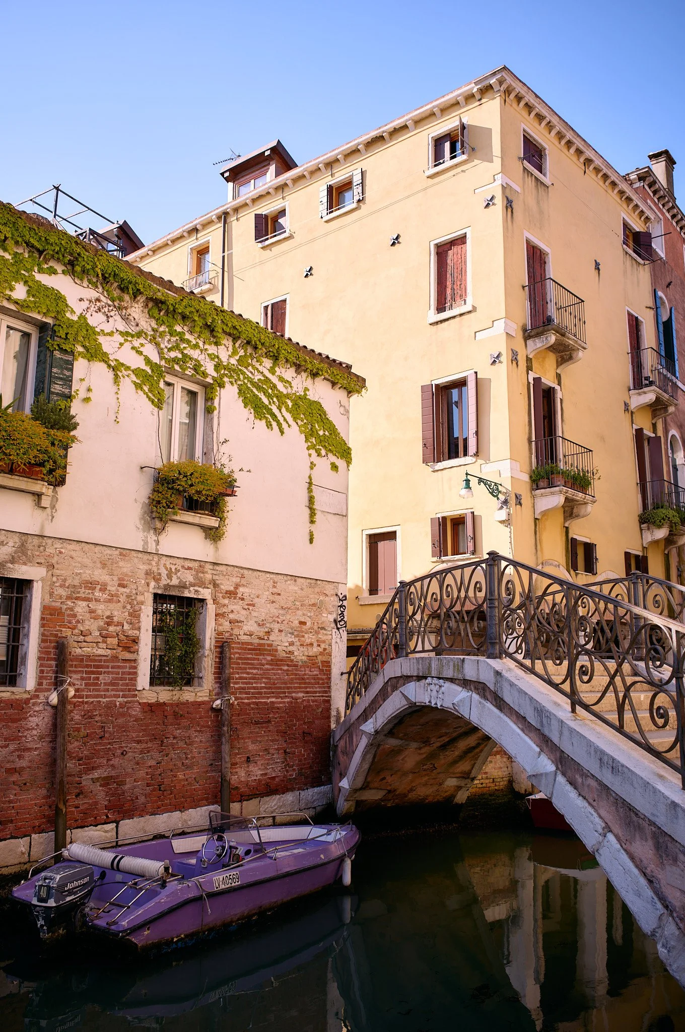 A small purple boat docked beside a canal, with old yellow and pink buildings with balconies and shuttered windows, and a small stone bridge arching over the water in Venice, Italy.