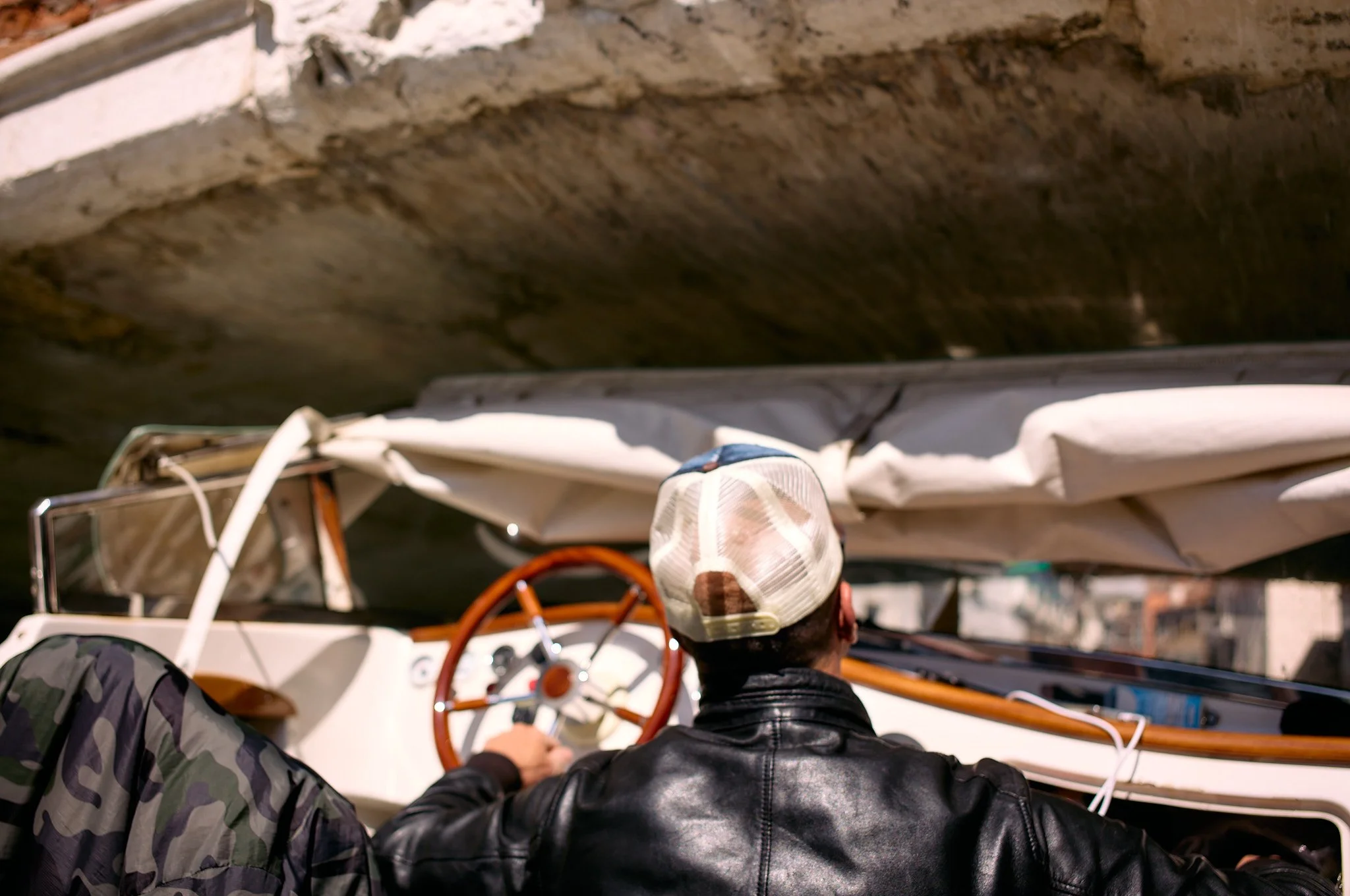 Person wearing a leather jacket and a camouflage cap sitting at the wheel of a small boat, with the boat under a bridge or tunnel.