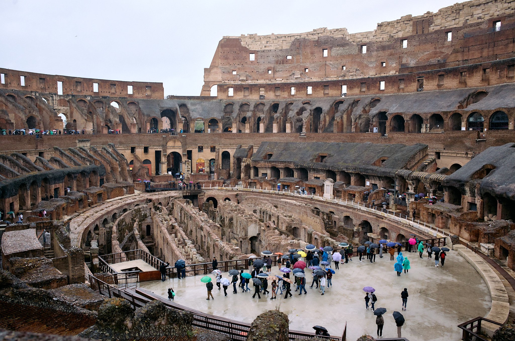 Tourists with umbrellas inside the ancient Roman Colosseum on a rainy day.
