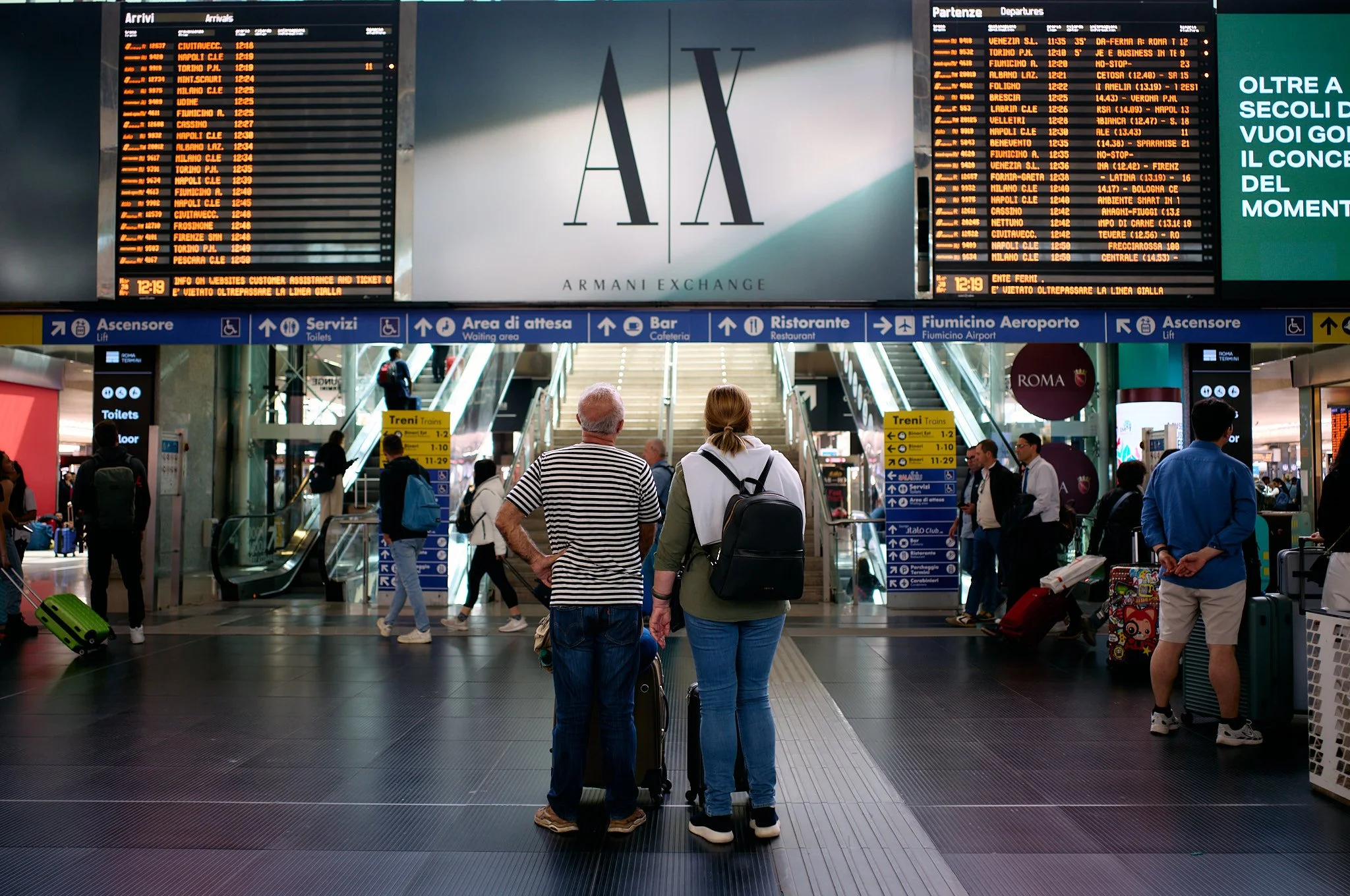 Passengers waiting at Fiumicino Airport terminal near escalators with departure and arrival boards overhead displaying flight information. The airport features signs for services such as toilets, elevators, waiting areas, bars, and restaurants. Two p