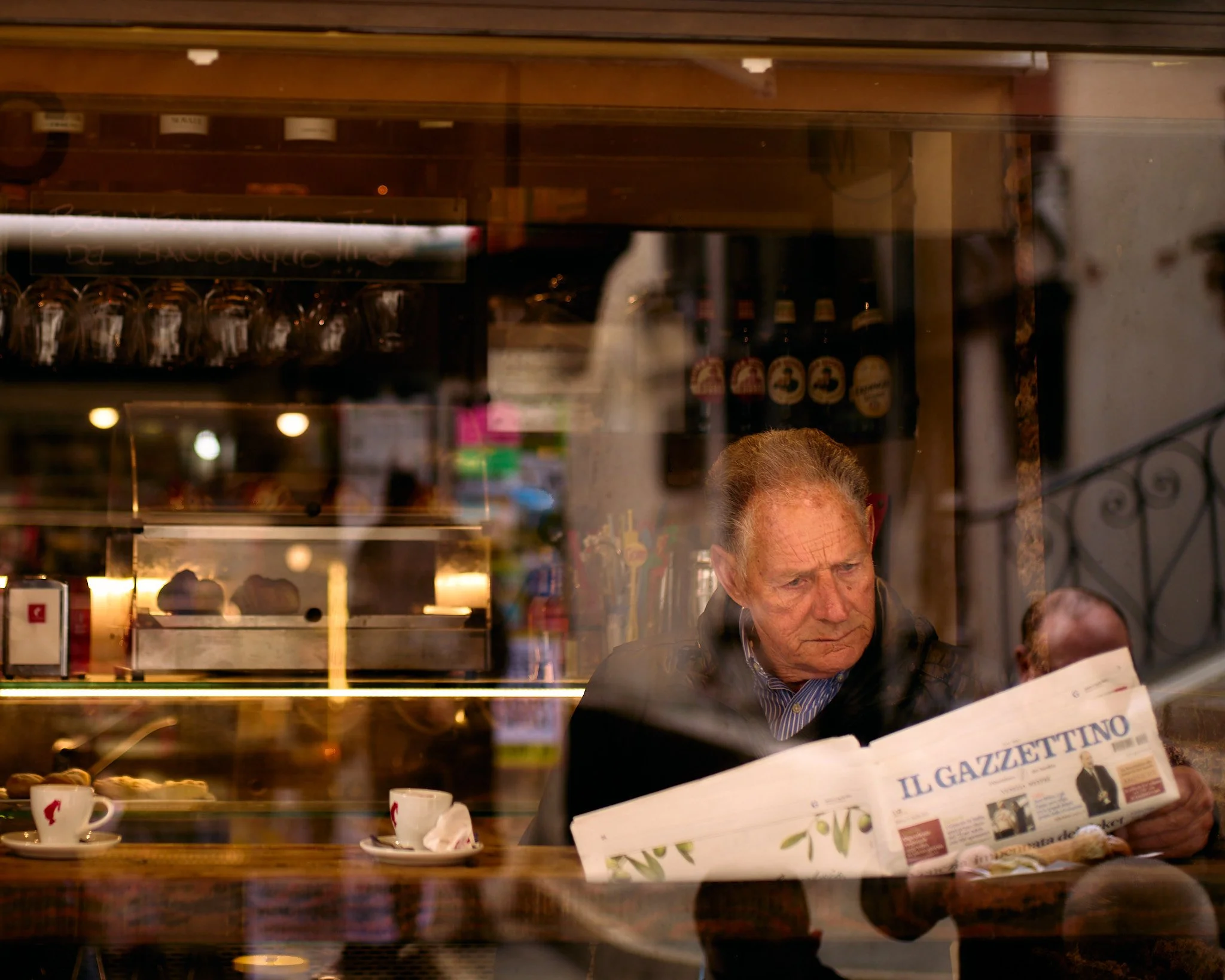 An older man is sitting inside a café, reading a newspaper titled 'IL GAZZETTINO,' through a window. The scene shows a wooden counter with cups and pastries, and the background displays shelves with bottles and glasses.