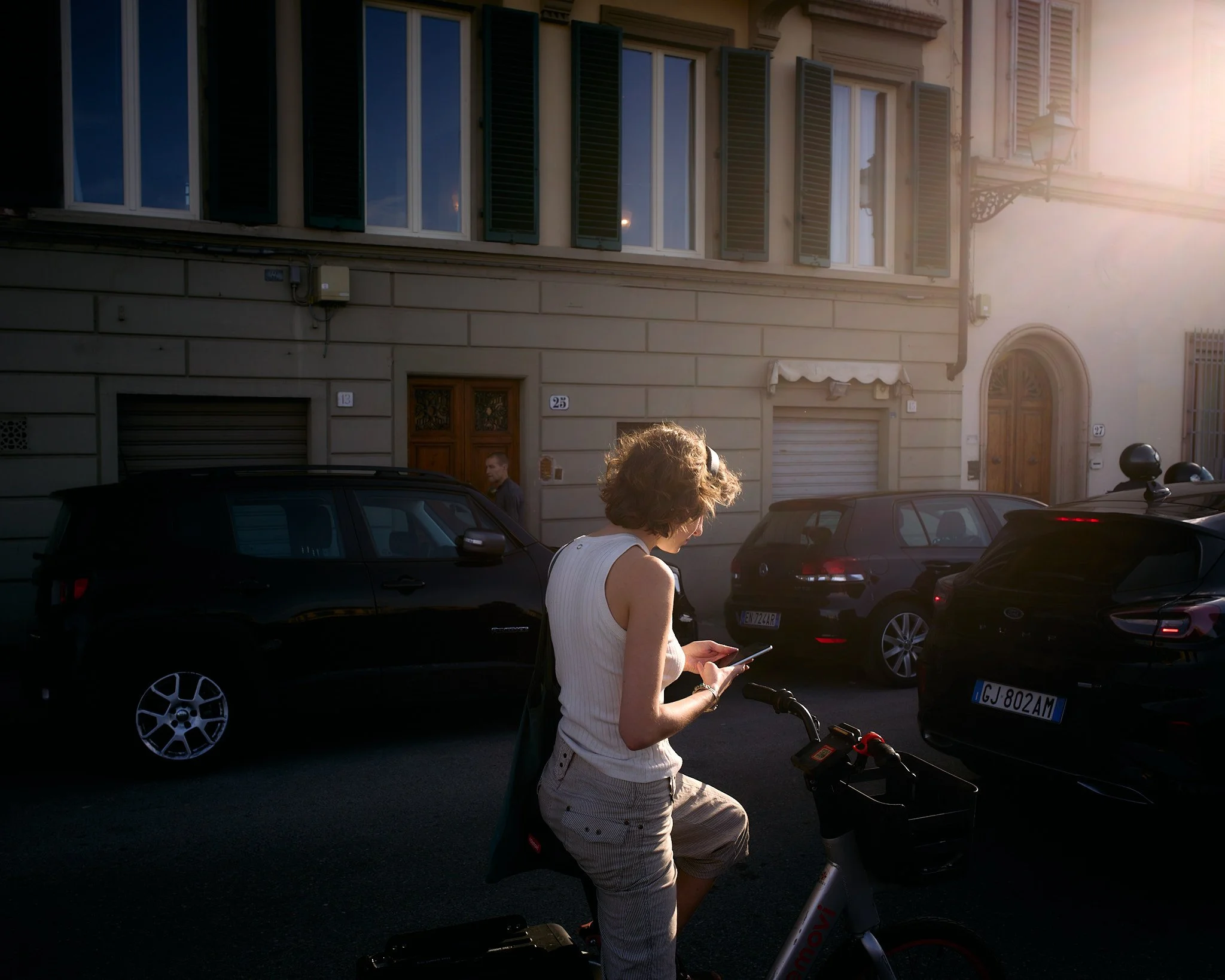 A woman in a white sleeveless top and striped pants stands with one foot on a bicycle, looking at her phone, on a city street with parked cars and old buildings in the background, illuminated by warm sunlight.
