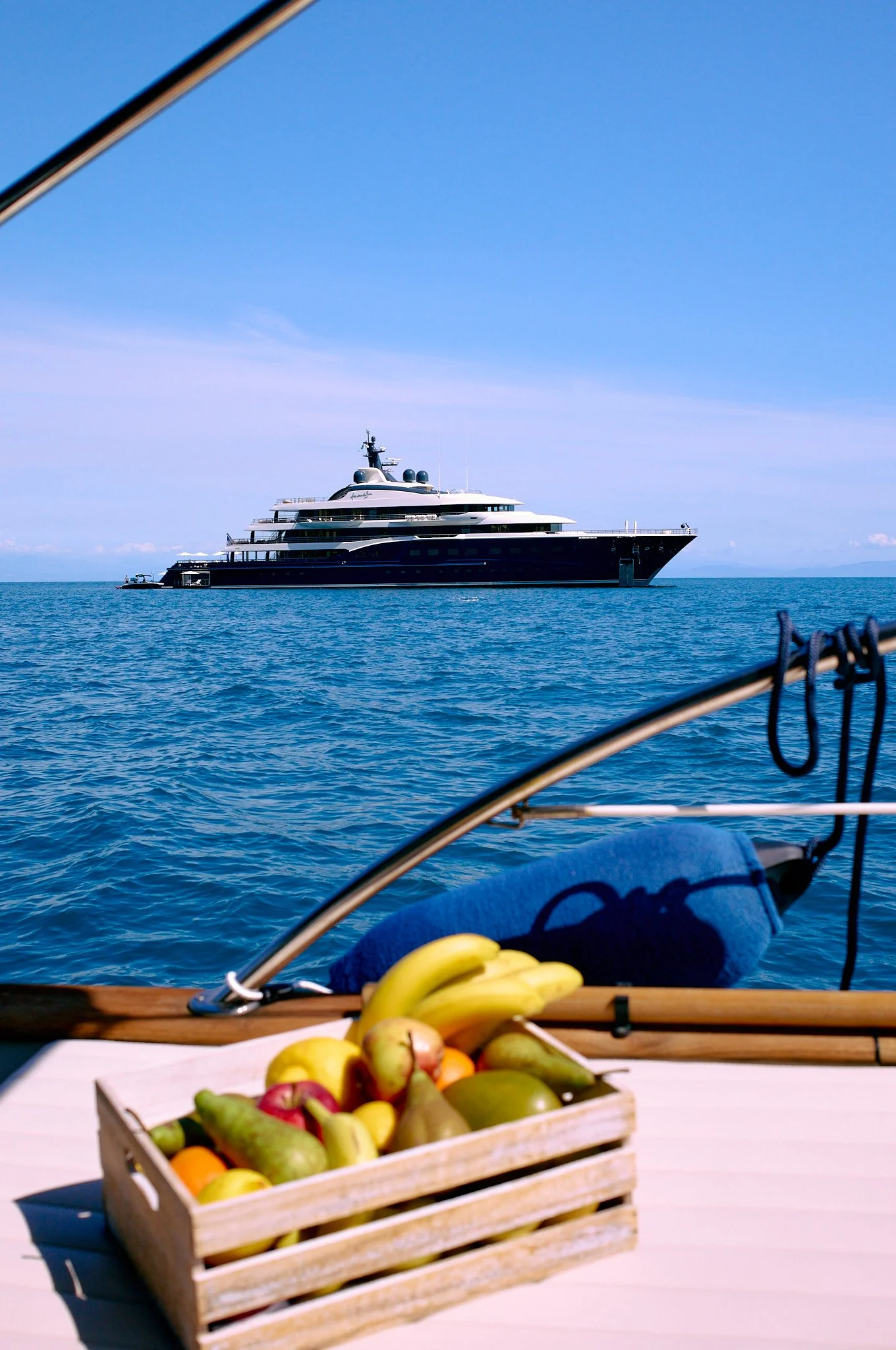 A wooden table on a boat with a basket of assorted fresh fruits, overlooking the sea with a large yacht in the distance under a clear blue sky.