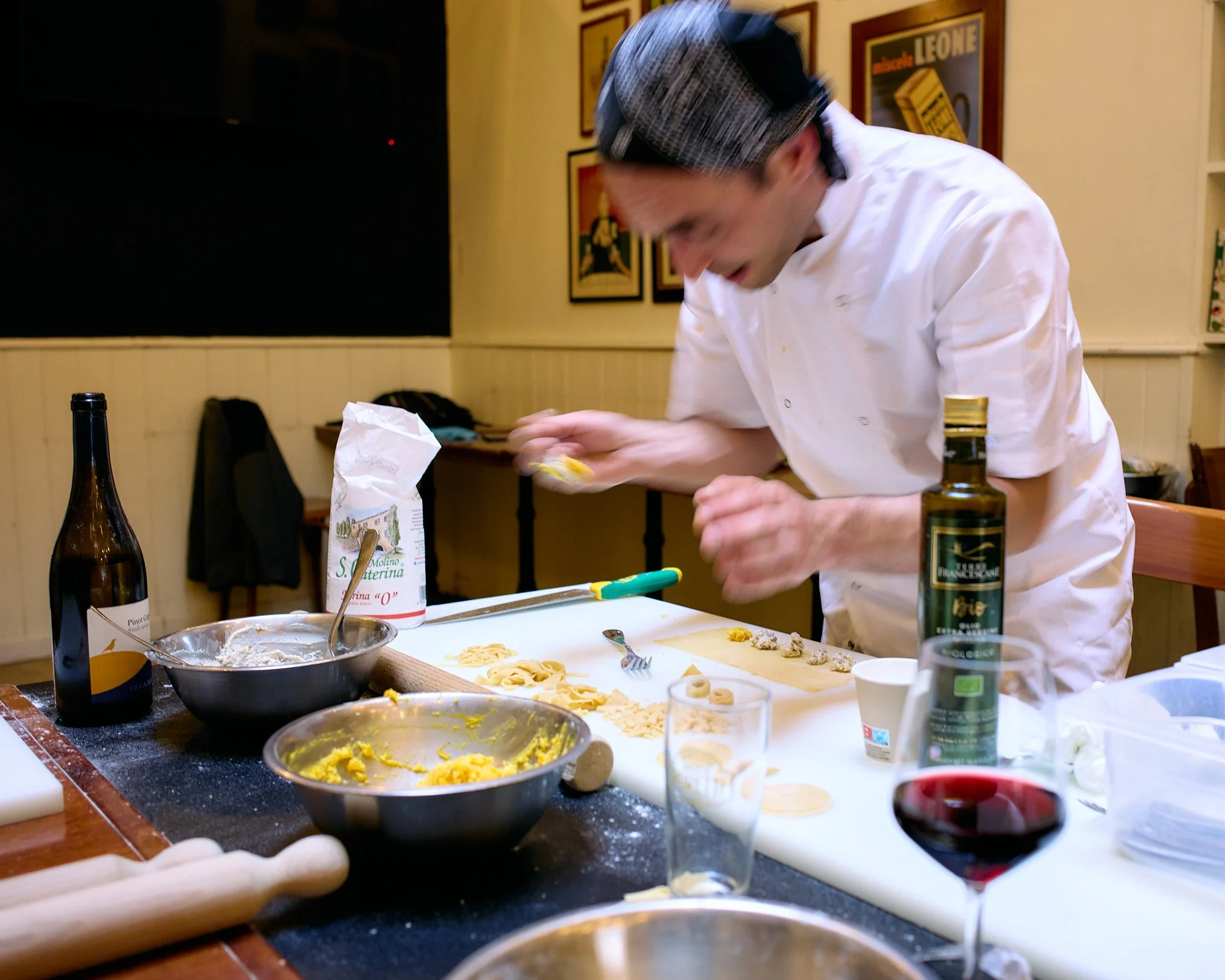 Chef in white uniform and black cap preparing pasta in a cozy restaurant kitchen, surrounded by cooking ingredients and utensils, with a glass of red wine in the foreground.