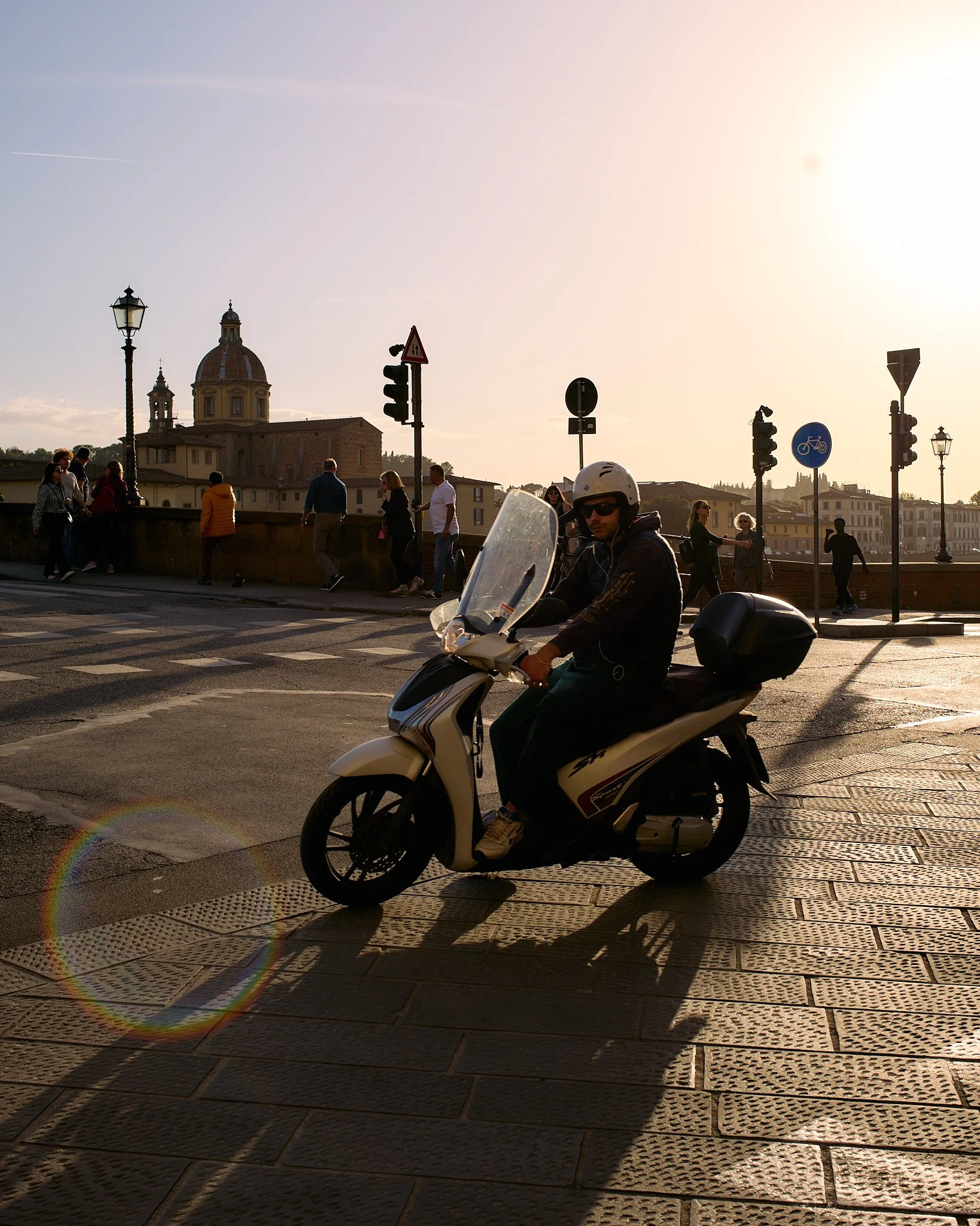 A man riding a white scooter on a city street during sunset, with pedestrians walking on the bridge and historic buildings in the background.
