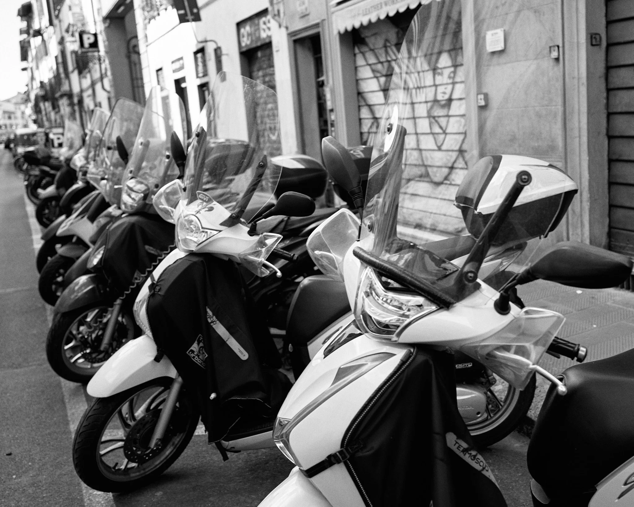 Several parked scooters with windshields lined up along a city street, storefronts, and buildings in the background, in black and white.