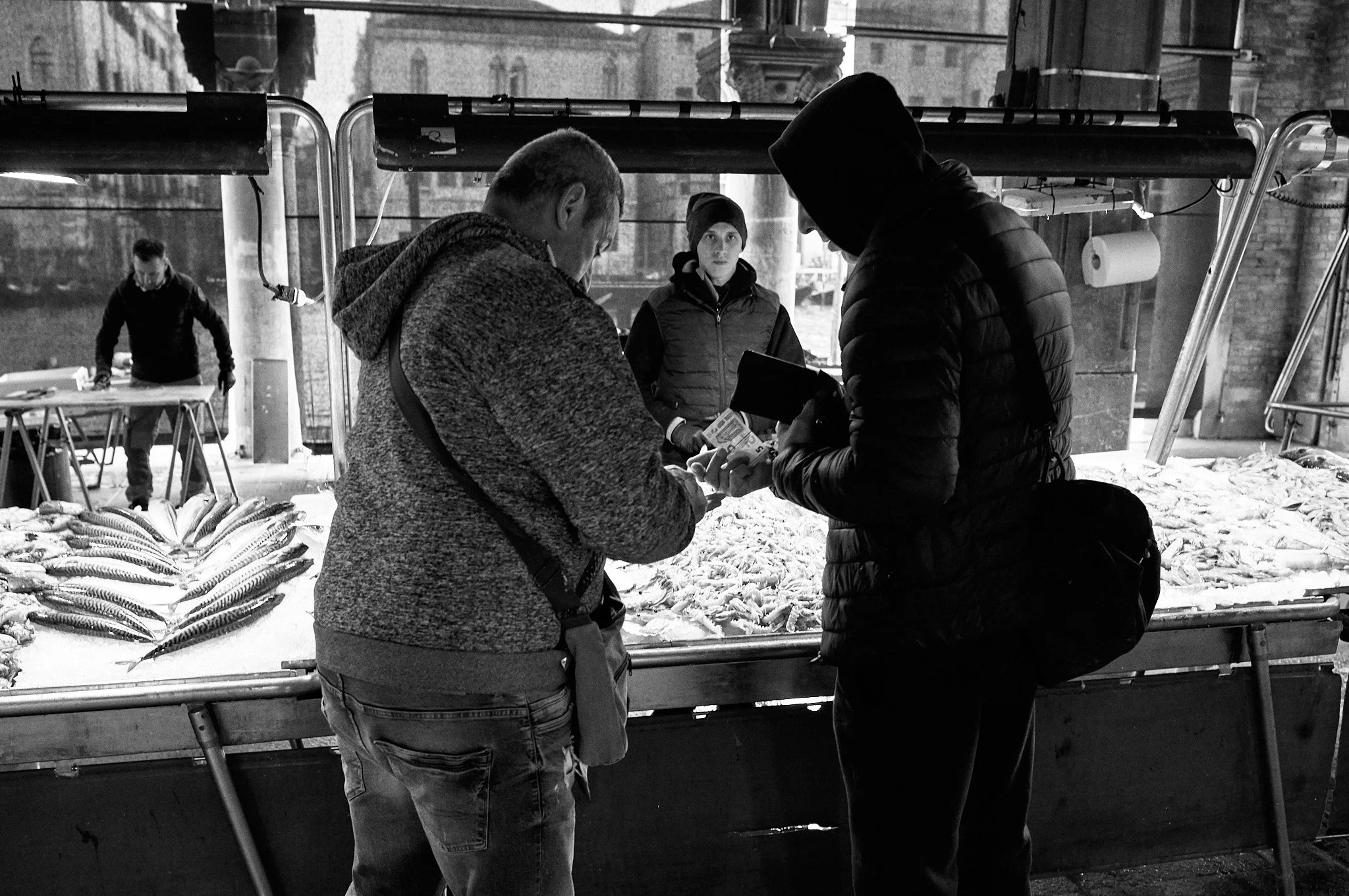 Two men are at a fish market counter, one is counting money and the other is holding a wallet or phone, with fish displayed on the counter. A woman stands behind them, looking at the transaction, with other vendors and customers in the background.
