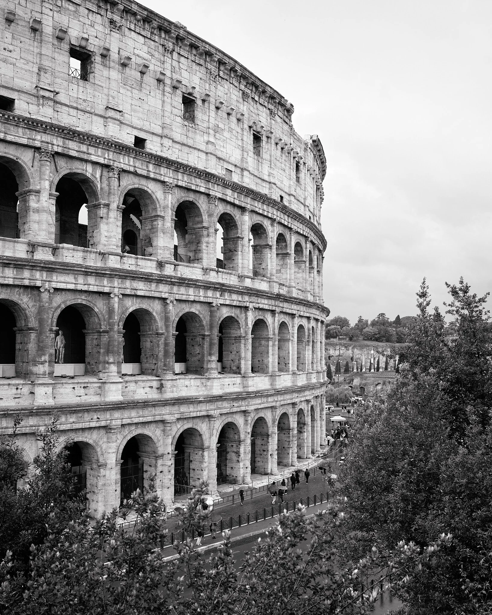 Black and white photo of the Colosseum in Rome, Italy, showing multiple levels of arches with people walking nearby and trees in the foreground.