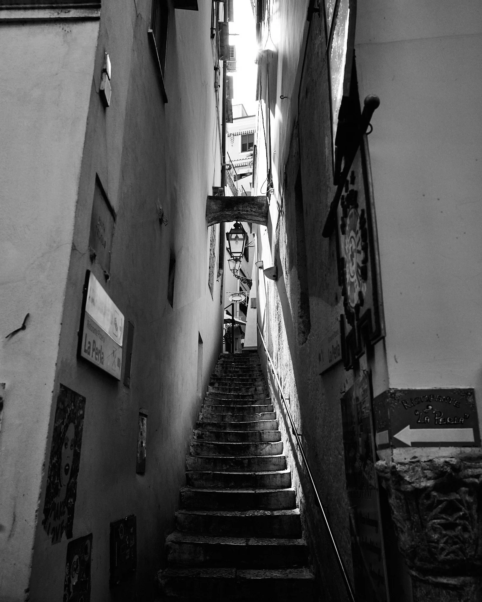 Narrow outdoor stairway between tall buildings, with lamps hanging and signs on the walls, leading to an open daylight street.
