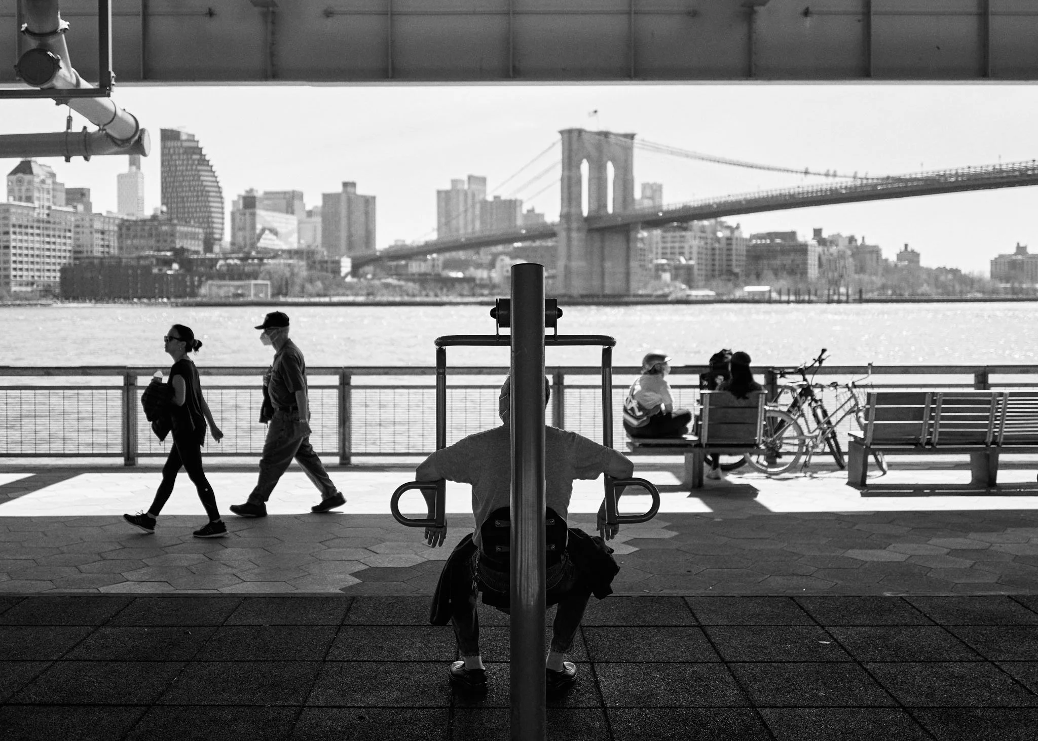 Black and white photo of a waterfront park with people walking and sitting, the Brooklyn Bridge and Manhattan skyline in the background.