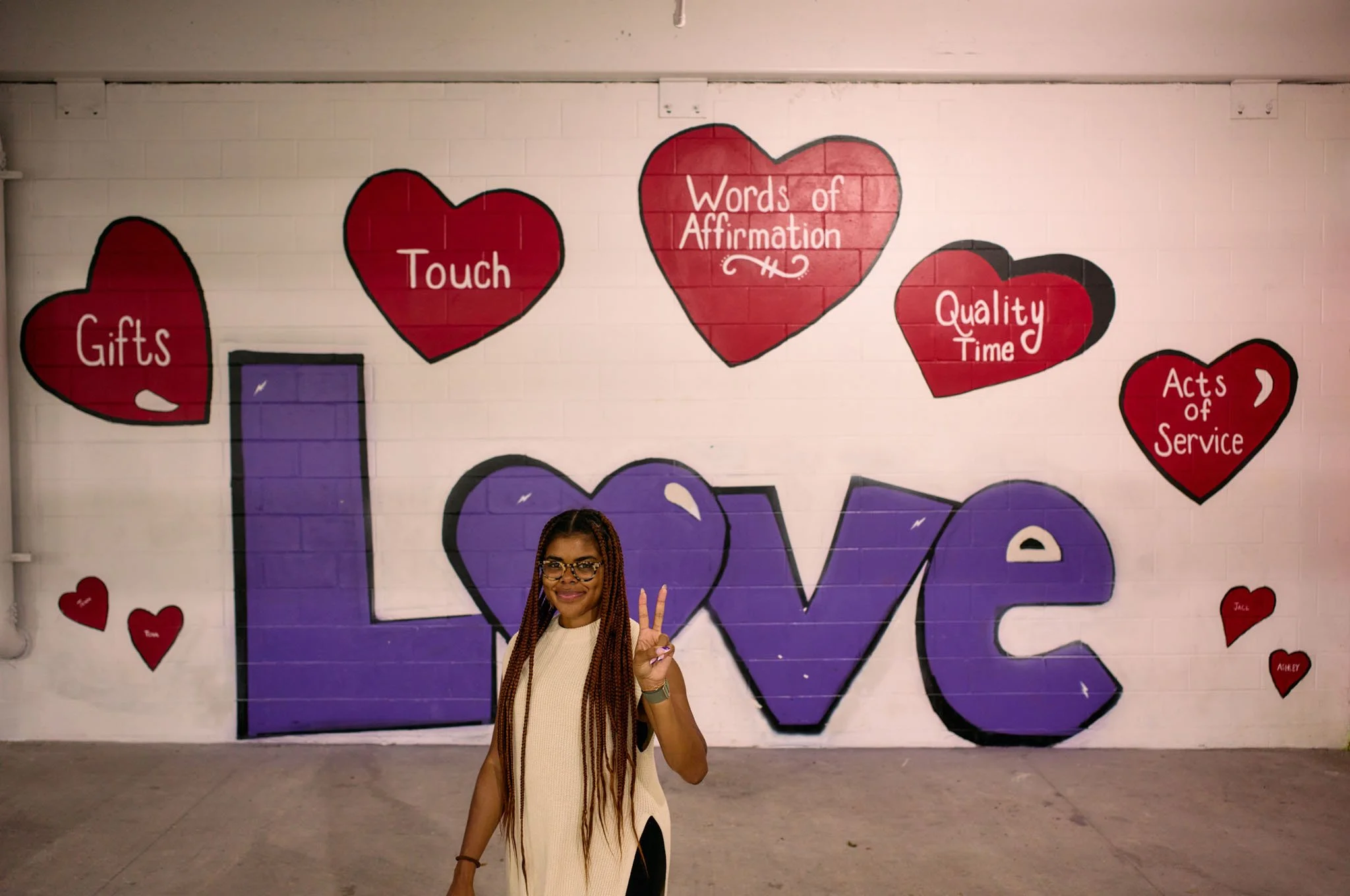 A woman with long braids and glasses standing in front of a wall with graffiti-style artwork spelling 'LOVE' in purple, surrounded by red hearts with words like 'Gifts,' 'Touch,' 'Words of Affirmation,' 'Quality Time,' and 'Acts of Service.'