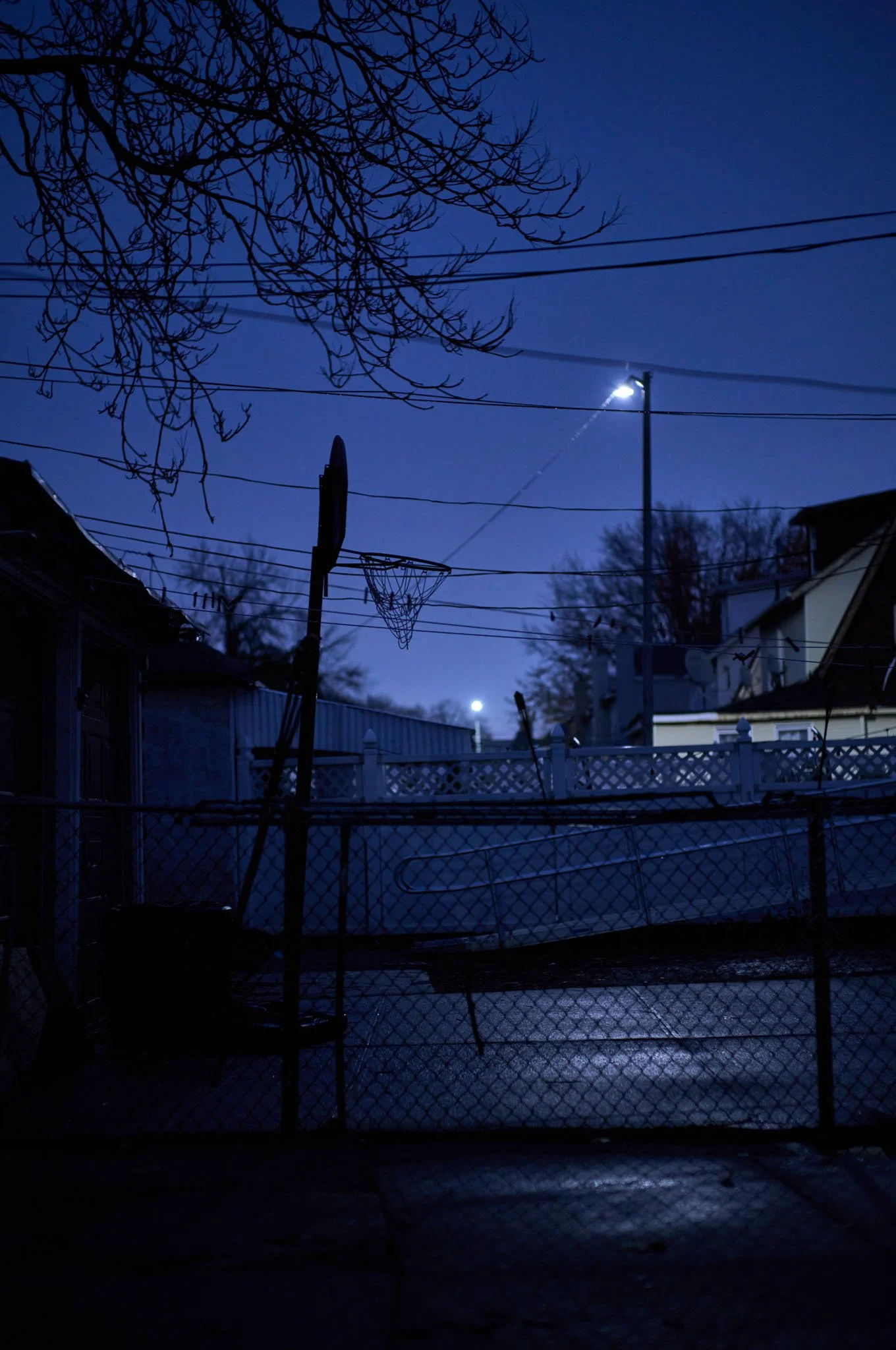 Nighttime scene of a backyard with a chain-link fence, a basketball hoop, power lines, bare tree branches, and a dark sky illuminated by distant lights.