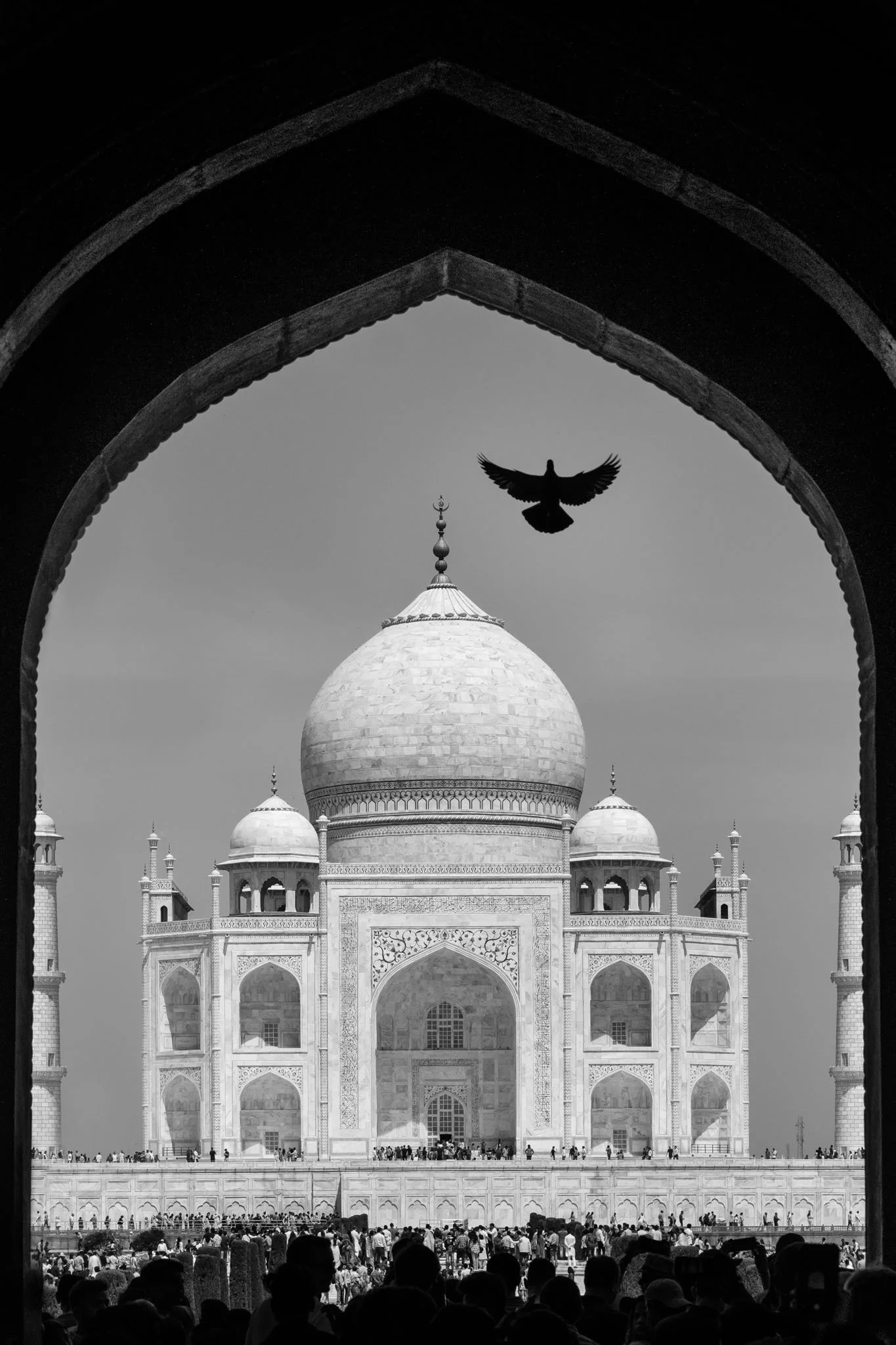 Black and white photo of the Taj Mahal with a large crowd of visitors in front, seen from a distance through an archway.