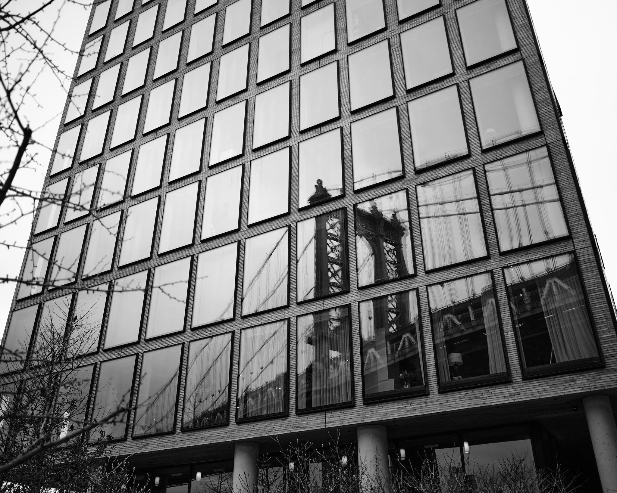 Reflection of the bridge tower in the glass windows of a modern multi-story building, with some trees in the foreground.