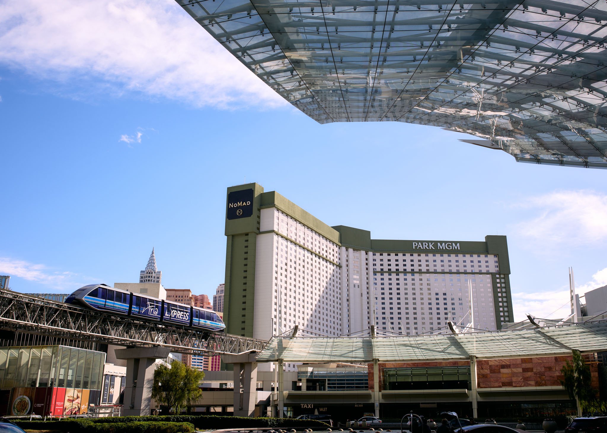 View of the Las Vegas Strip with the MGM Grand hotel, a monorail train labeled 'ARIA Express,' modern buildings, and a blue sky with some clouds.