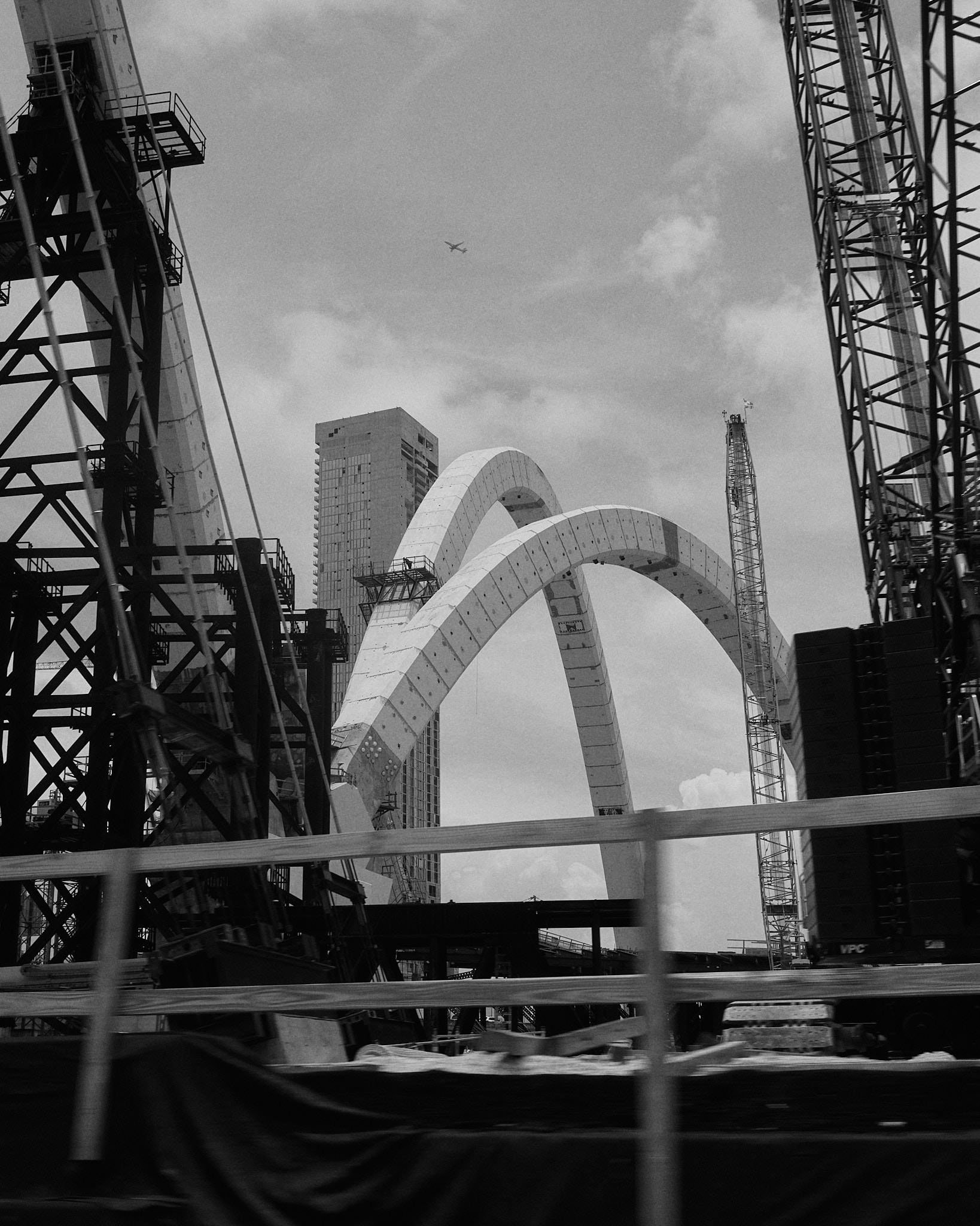 Construction site with large arches, cranes, and high-rise buildings, with an airplane flying in the sky.