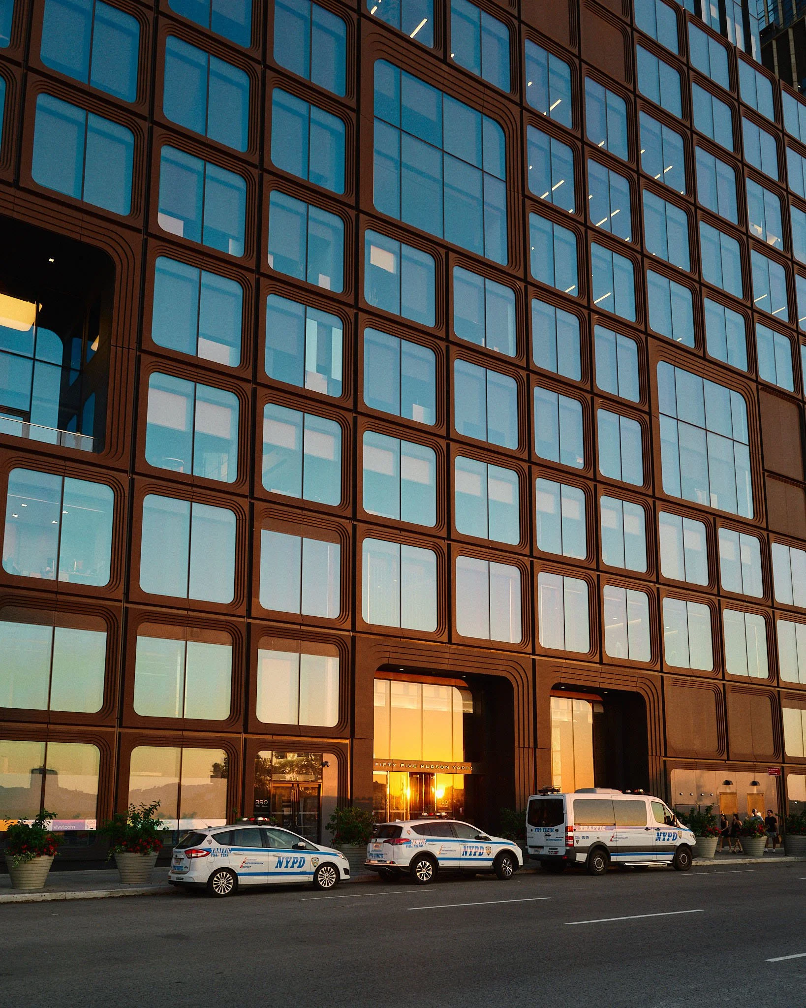 City building with reflective glass windows and NYPD police vehicles parked in front at sunset.