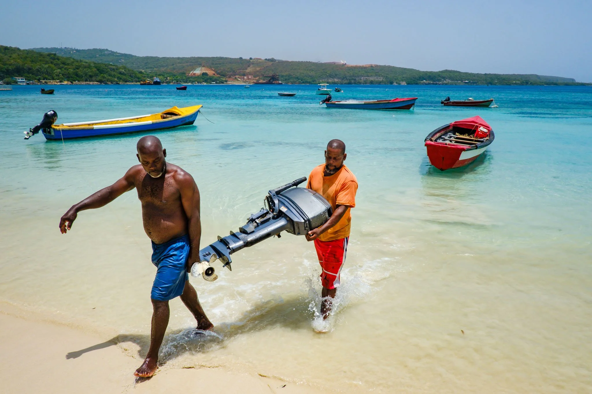 Two men with dark skin walking out of the water on a sandy beach, one shirtless and the other wearing an orange t-shirt, carrying a boat motor. Several colorful boats float on clear blue water with a hilly landscape in the background.
