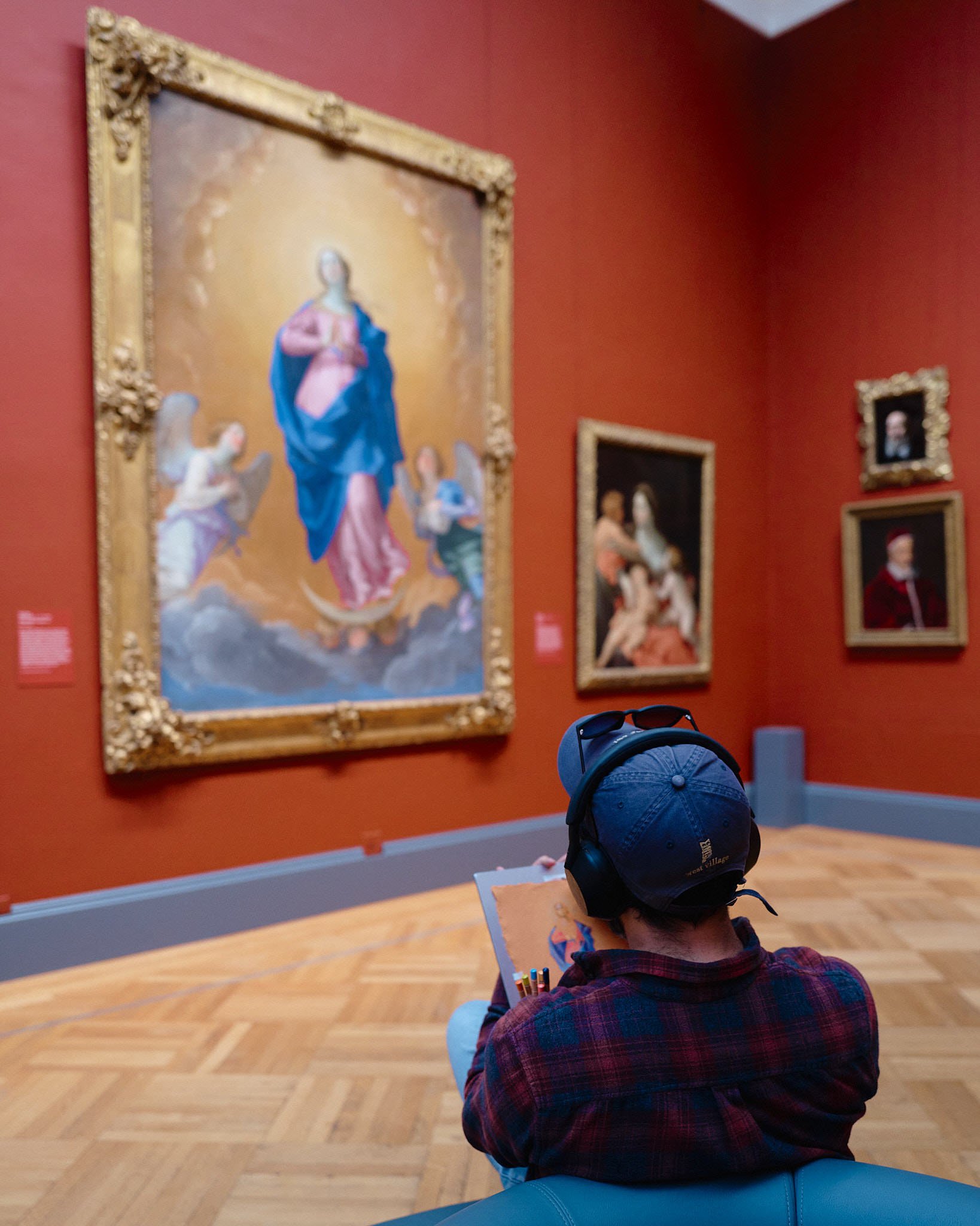 A person with a blue cap, headphones, and sunglasses resting on their head, sitting and drawing in a sketchbook while viewing paintings in an art gallery with red walls and wooden flooring.