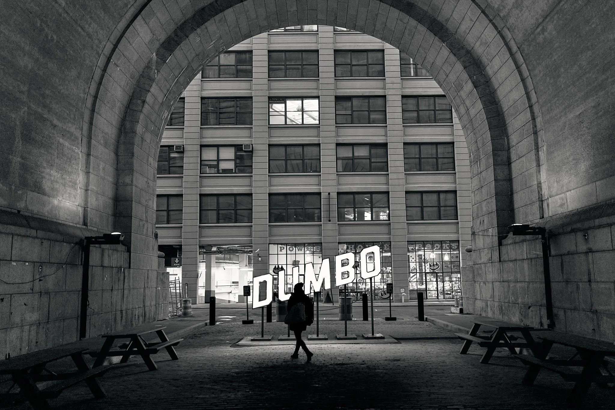A person walking through an archway, with a large illuminated sign spelling 'DUMBO' in front of a modern building with many windows.