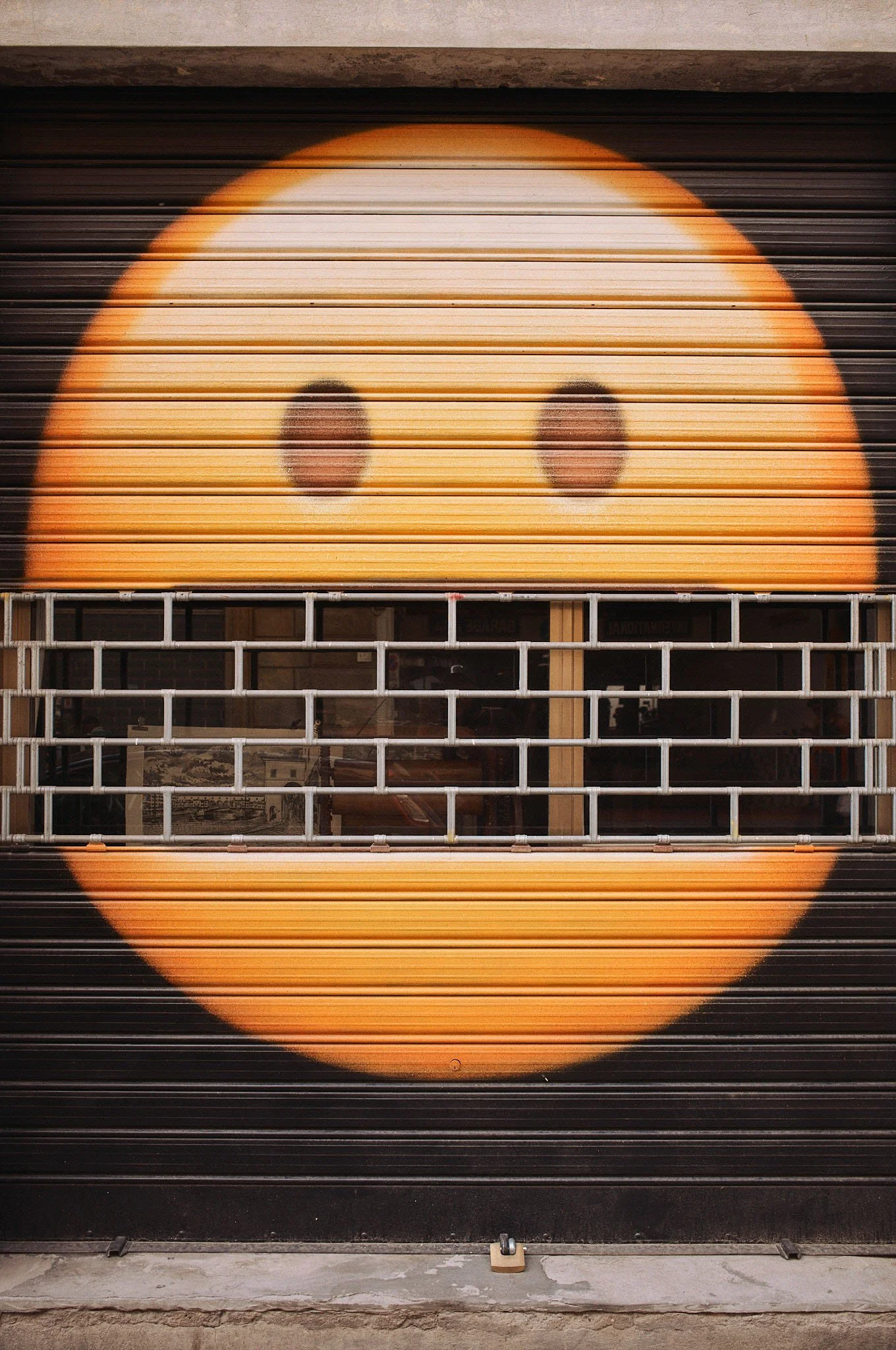 A yellow smiley face painted on a metal roller shutter with grid-like security bars across the middle.