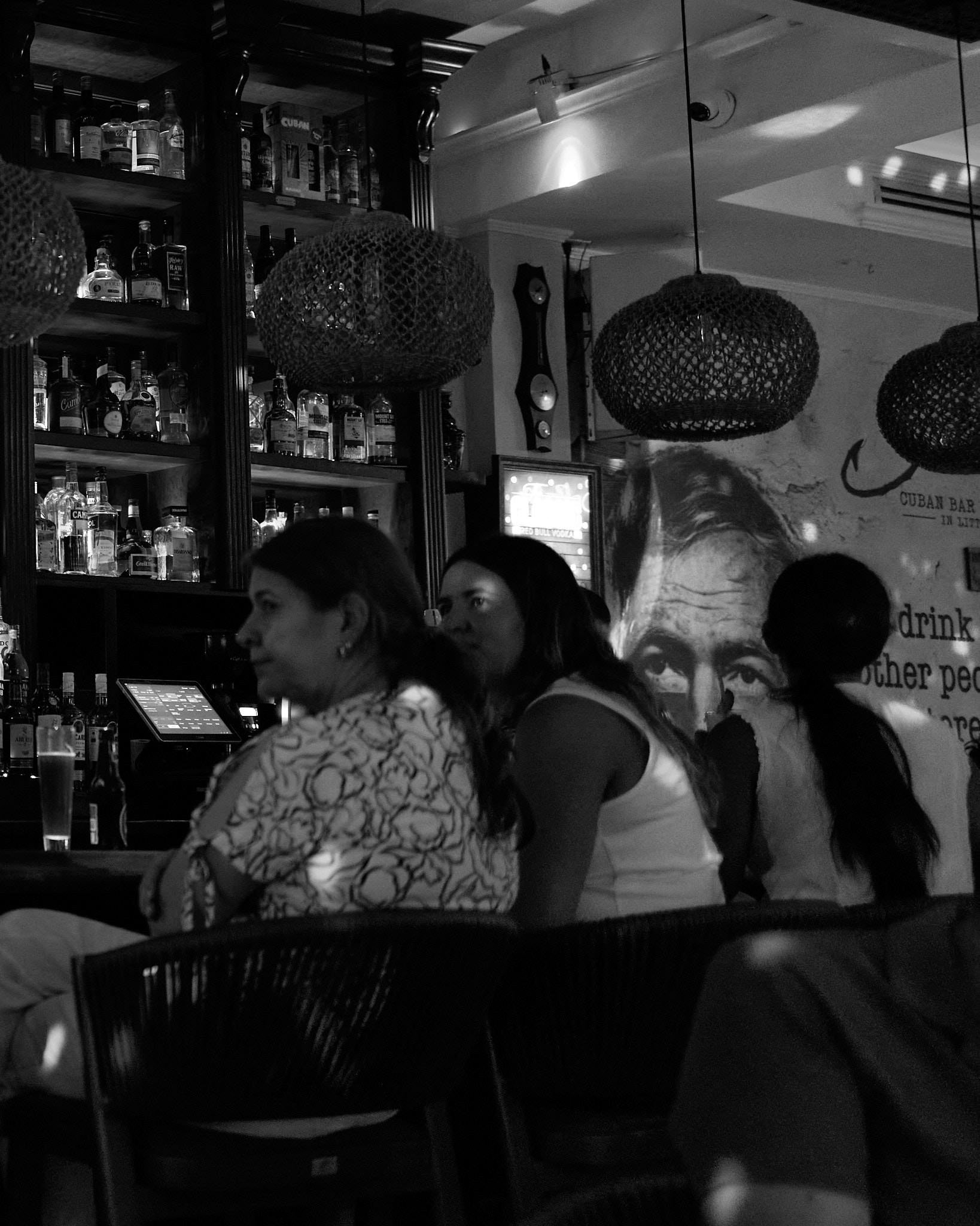 Three women sitting at a bar in a Cuban-themed bar, with bottles of alcohol on shelves behind them and hanging wicker lamps above.