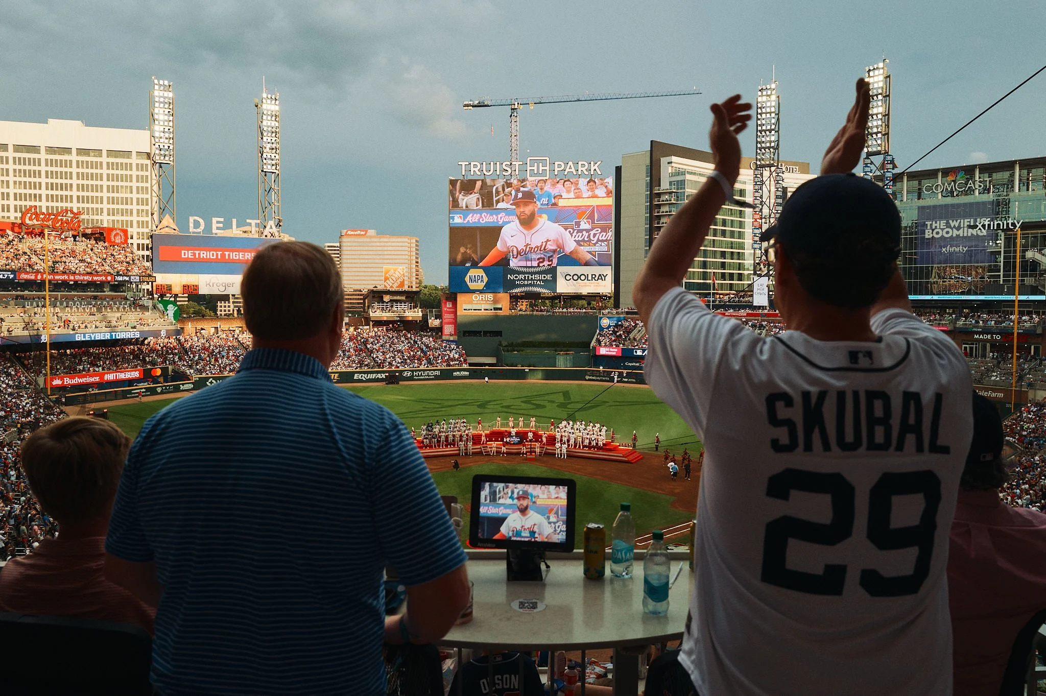 View of a baseball stadium during a game with spectators in the stands and a large electronic scoreboard displaying a player. Foreground shows fans, one wearing a San Diego Padres jersey with the name Skubal and the number 29, and others watching the