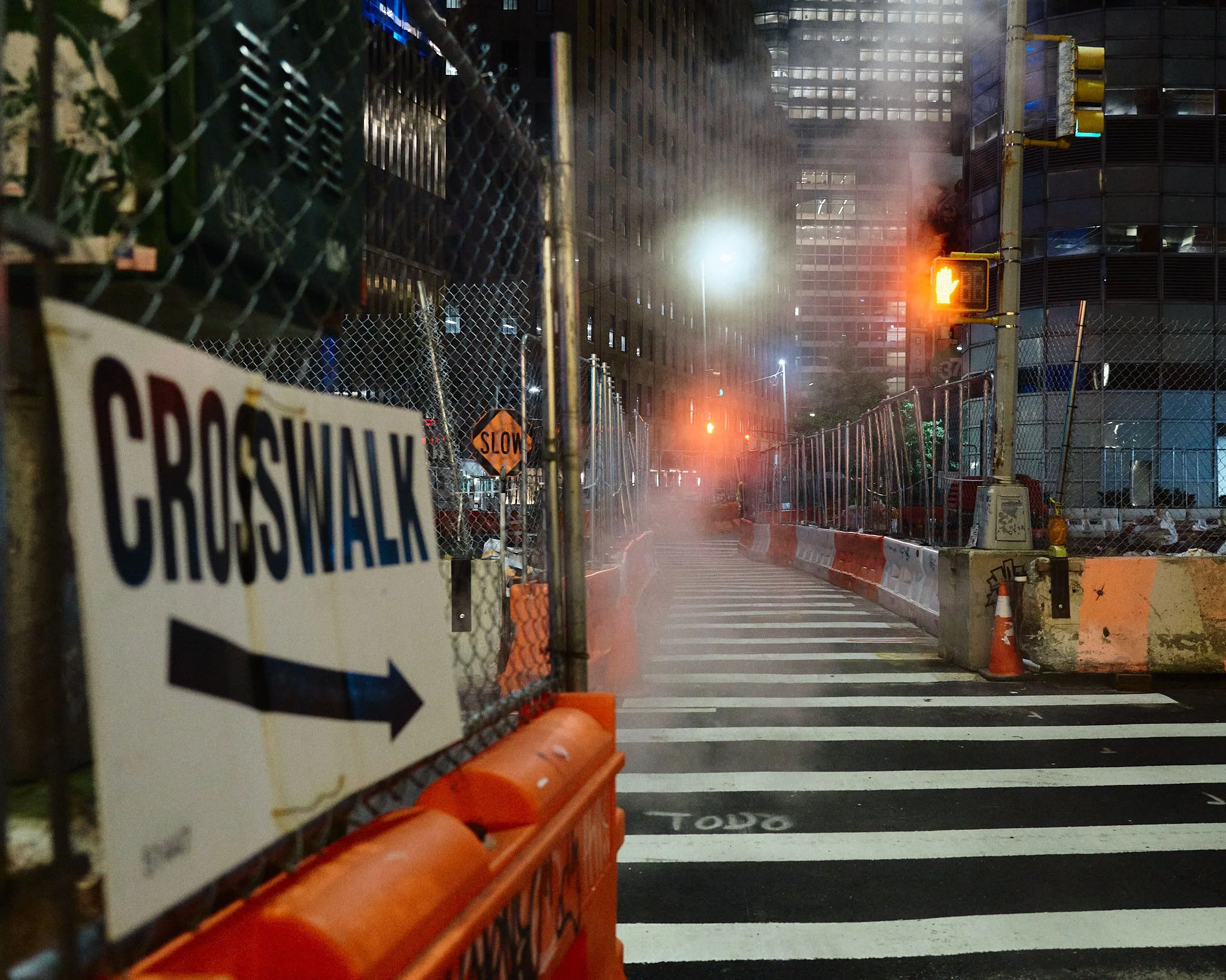 Nighttime city street under construction with a crosswalk, construction barriers, fencing, a 'CROSSWALK' sign with an arrow, and traffic lights showing a pedestrian signal and a yellow light. Smoke or steam is visible in the background.