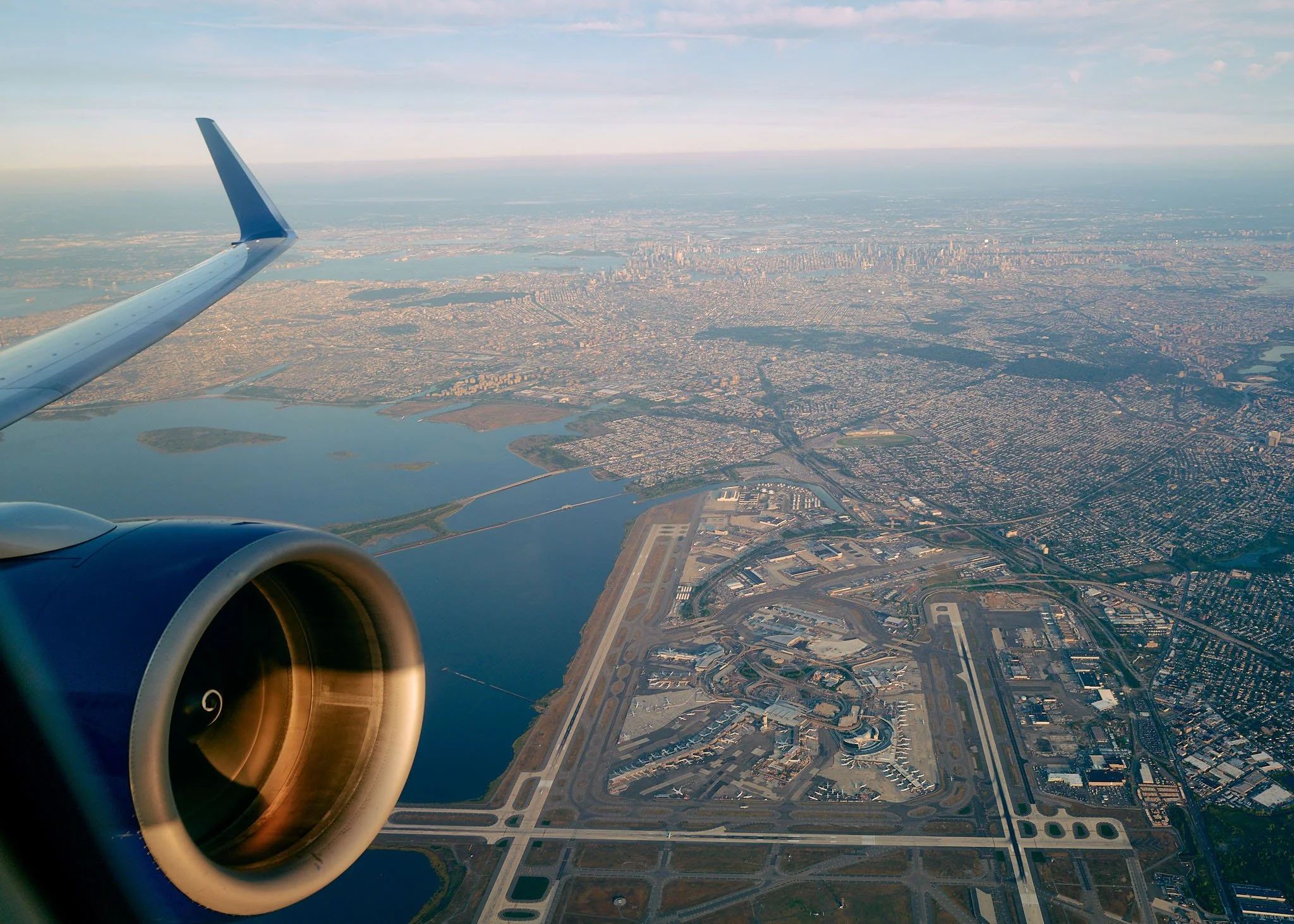 Aerial view of a city seen from an airplane window with a visible engine and wing, showing urban landscape, water bodies, and a runway.