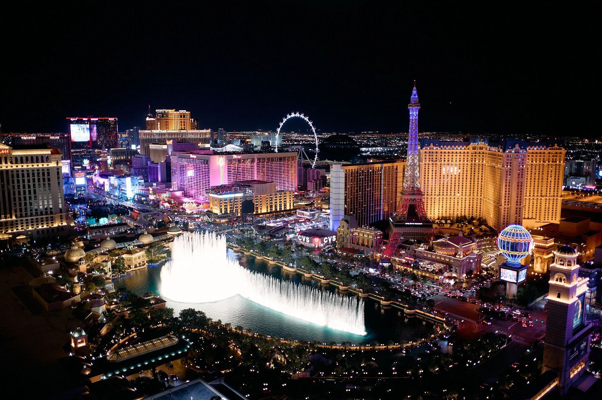 Night view of Las Vegas Strip with illuminated buildings, a large fountain display, the Eiffel Tower replica, and a Ferris wheel.