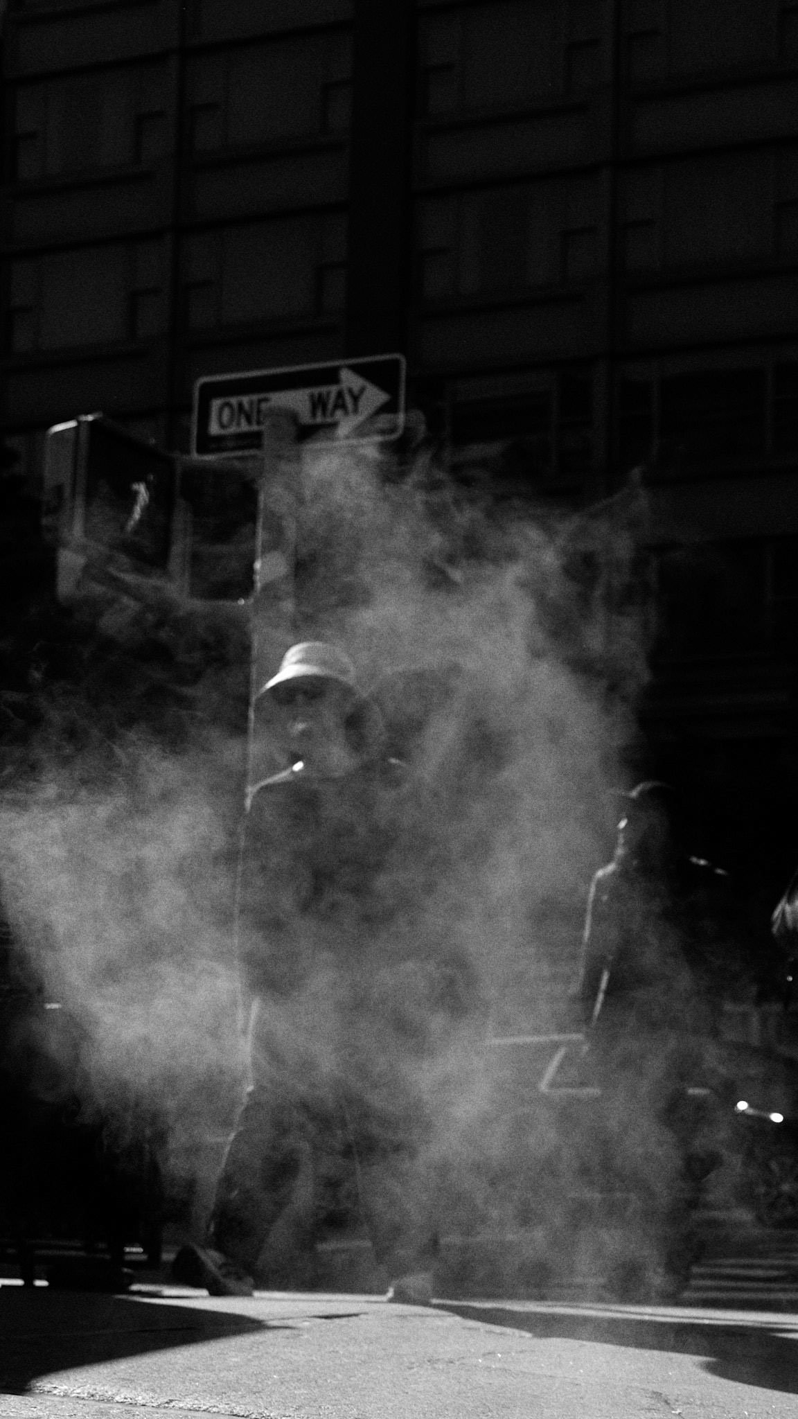 Two individuals walking in foggy urban street at night, one wearing a wide-brimmed hat and smoking.