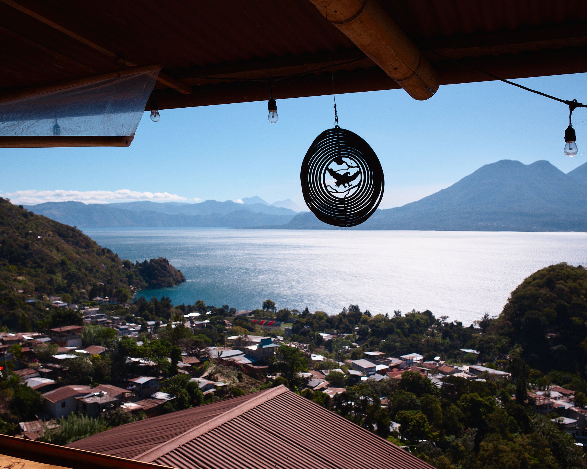 A scenic view of a lake surrounded by mountains, with a small town at the base, taken from a covered balcony with string lights and a decorative hanging ornament featuring a lizard inside concentric circles.