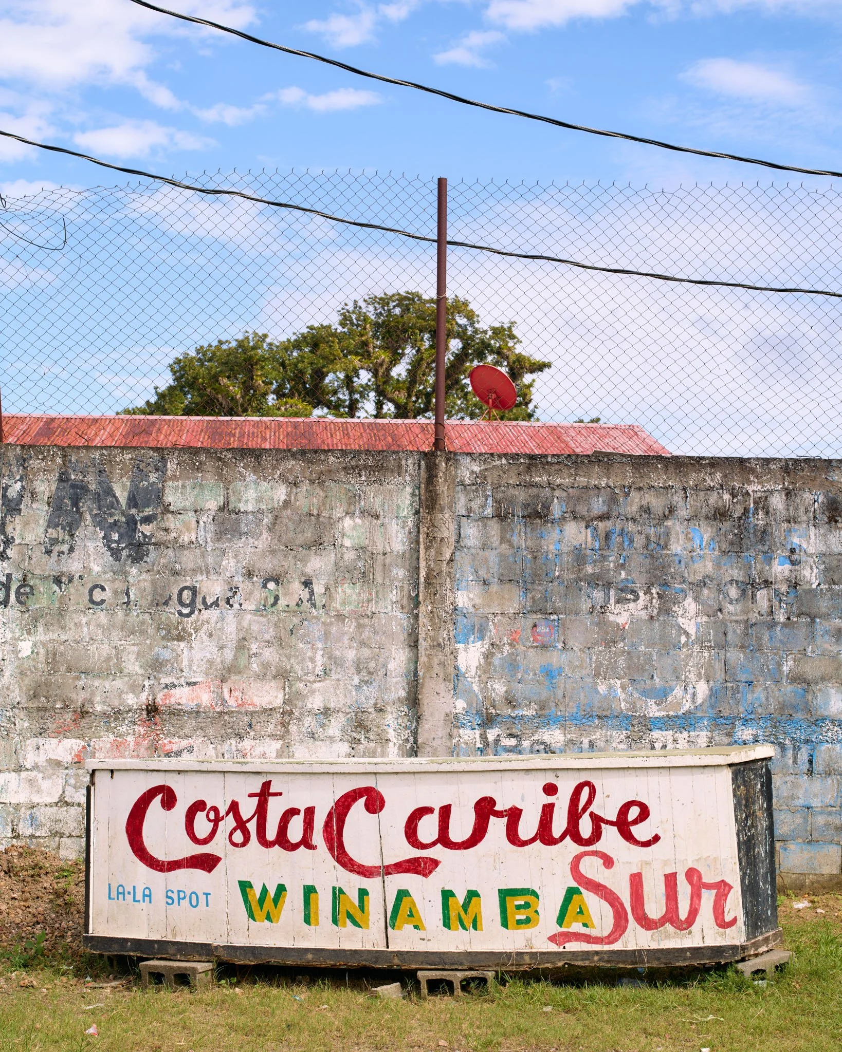 Colorful hand-painted sign on a white wooden board with the words "Costa Curibe, La-La Spot, Winamba, Sur." The background features a concrete wall with graffiti, a red-tiled roof, a satellite dish, and a blue sky with white clouds.