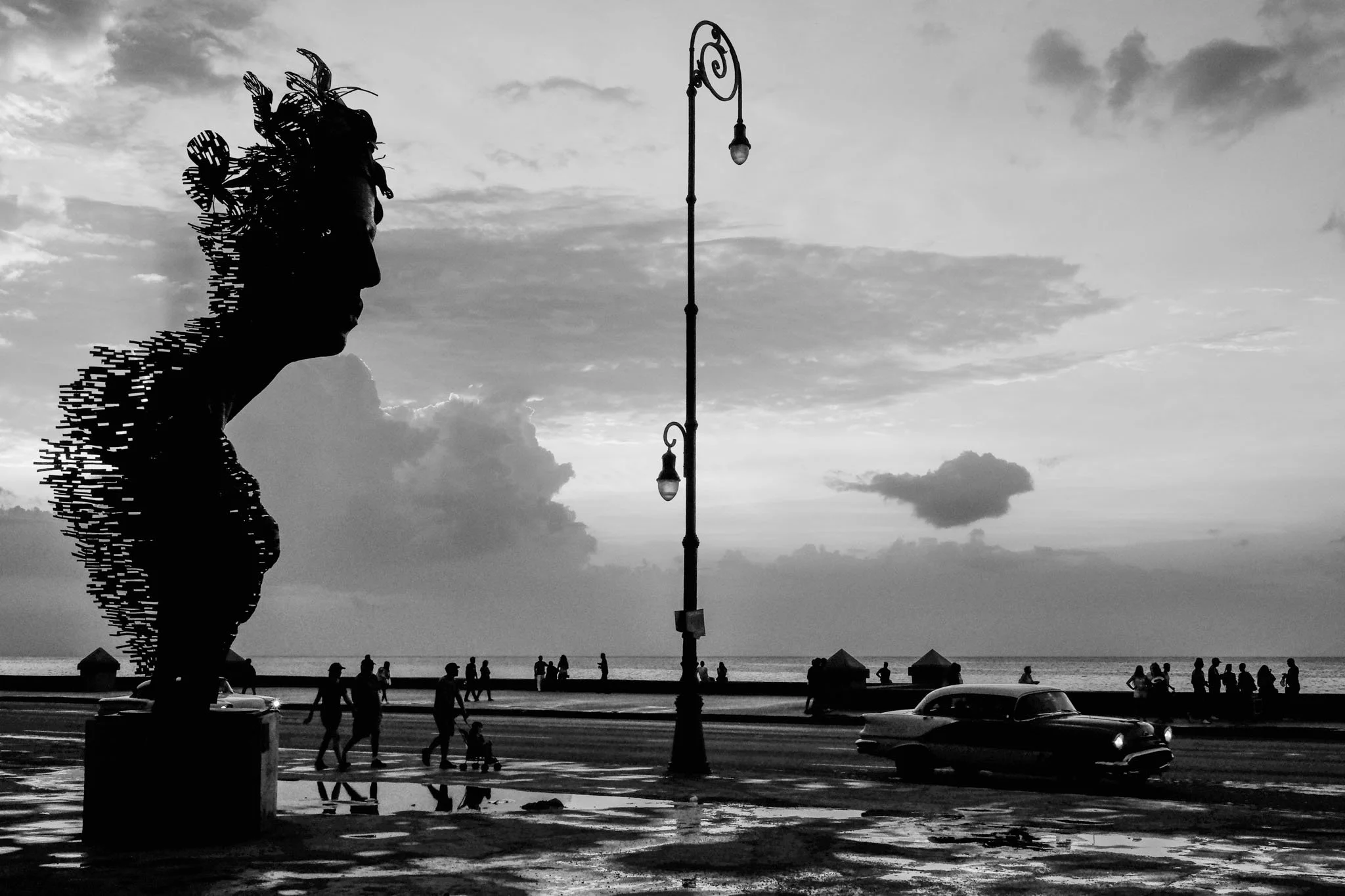 Black and white photo of a seaside promenade with a large sculpture of a woman's profile adorned with butterflies on the left. A tall street lamp is in the center, and people walking along the promenade can be seen in the background, with the ocean a