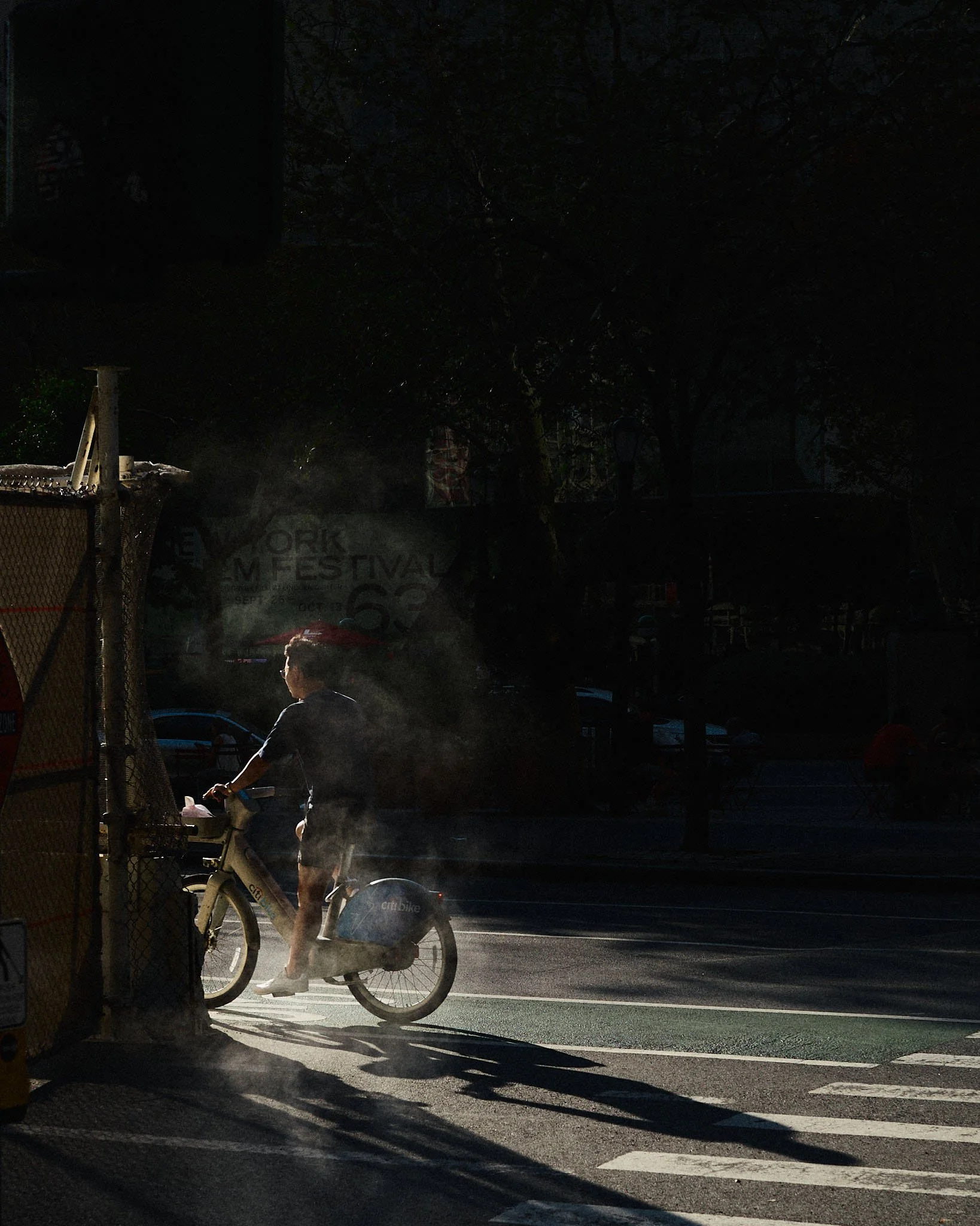 A person riding a bike at sunset or sunrise, with steam or smoke rising near the bike and crossing a street with a pedestrian crosswalk.