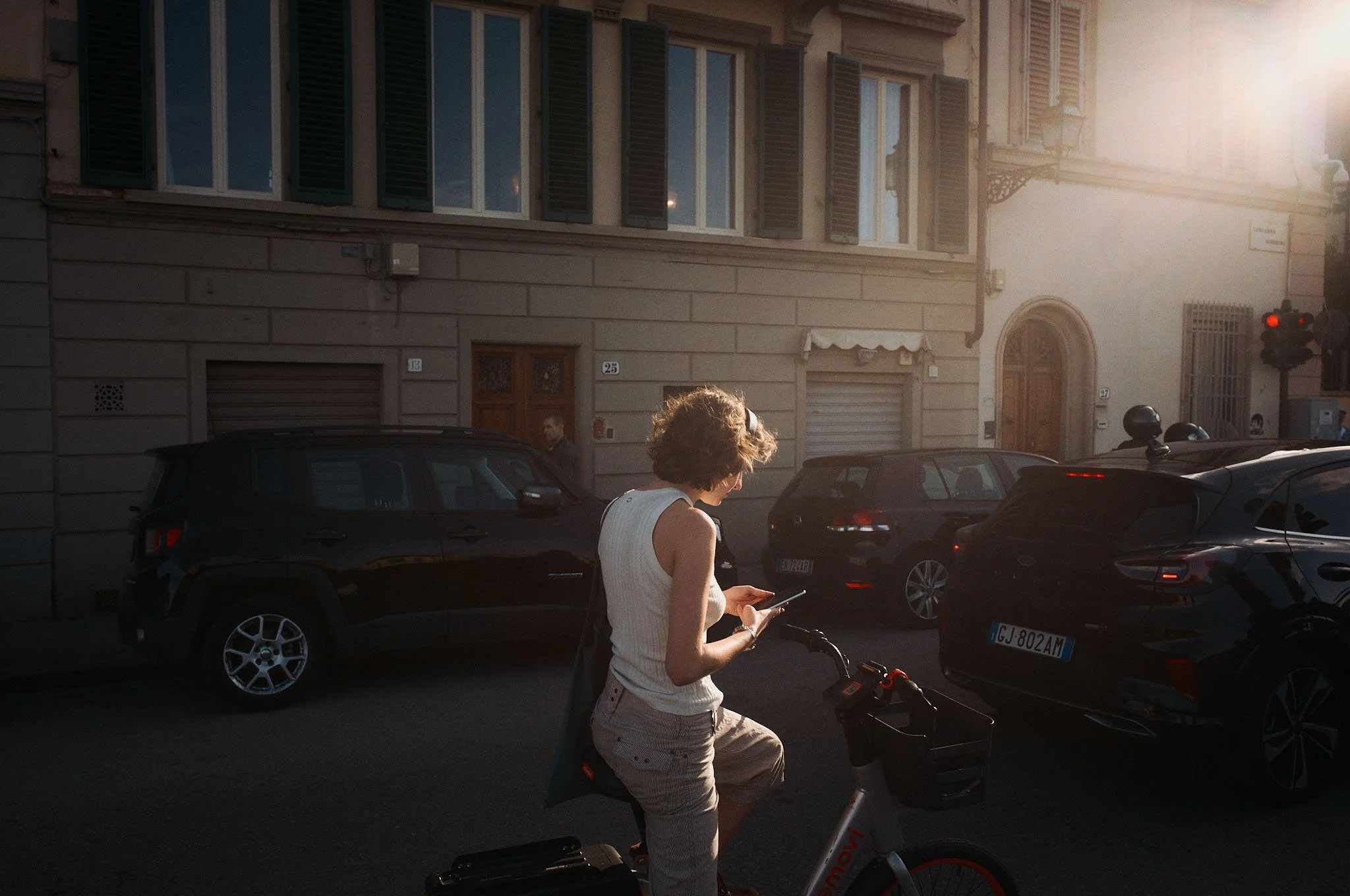 A woman stands on an electric bicycle using her phone in a city street during sunset, with parked cars and buildings in the background.